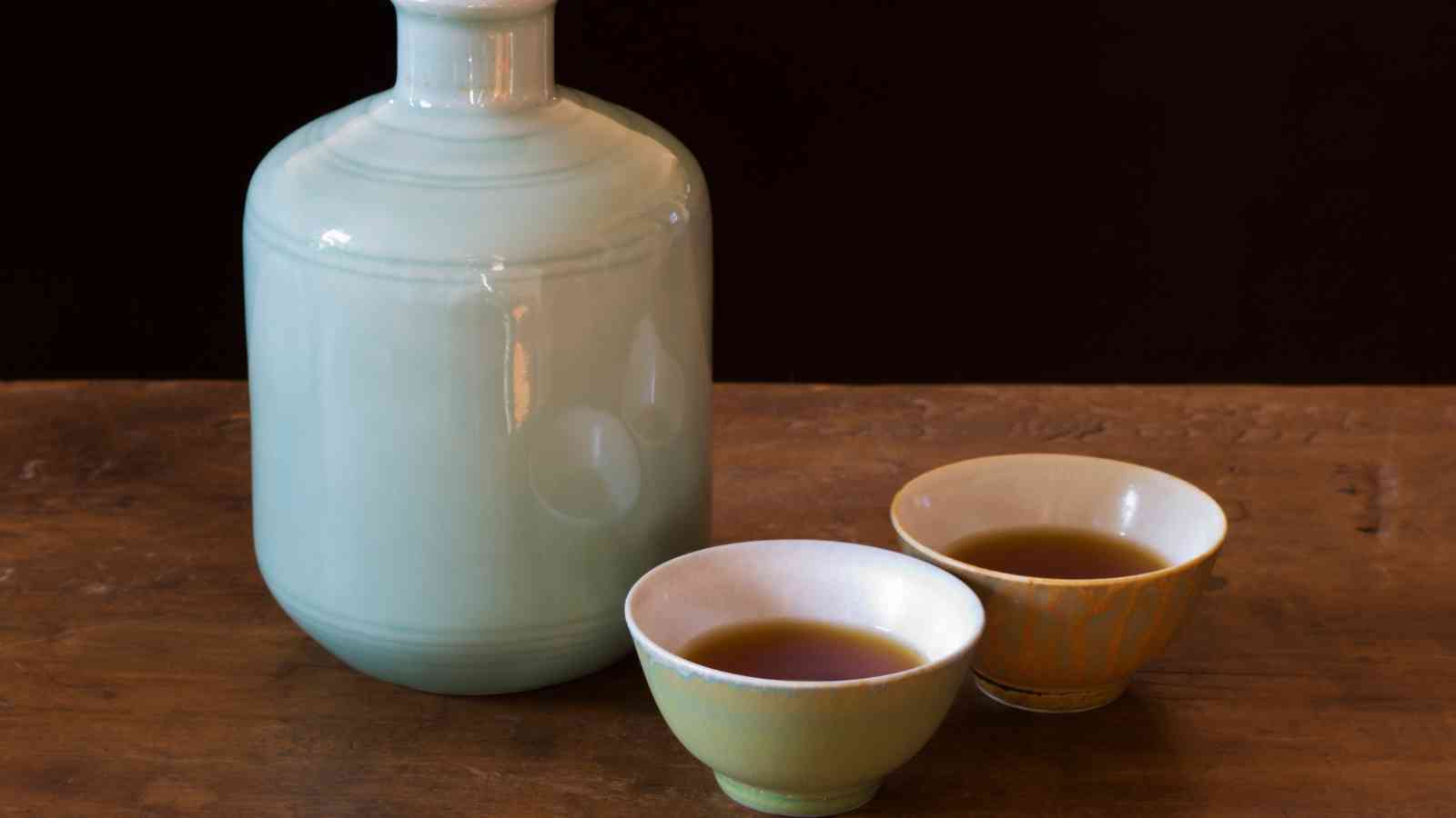 A pale ceramic Shaoxing wine bottle and two small cups filled with Shaoxing wine are placed on a wooden surface against a dark background.