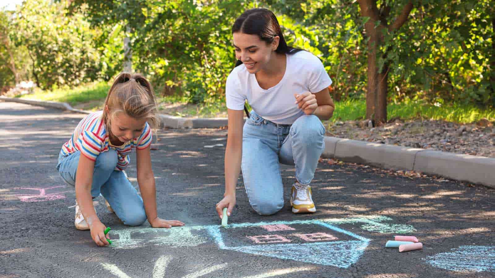 An adult and a child draw a house and other shapes with colored chalk on a paved path outdoors, surrounded by greenery.