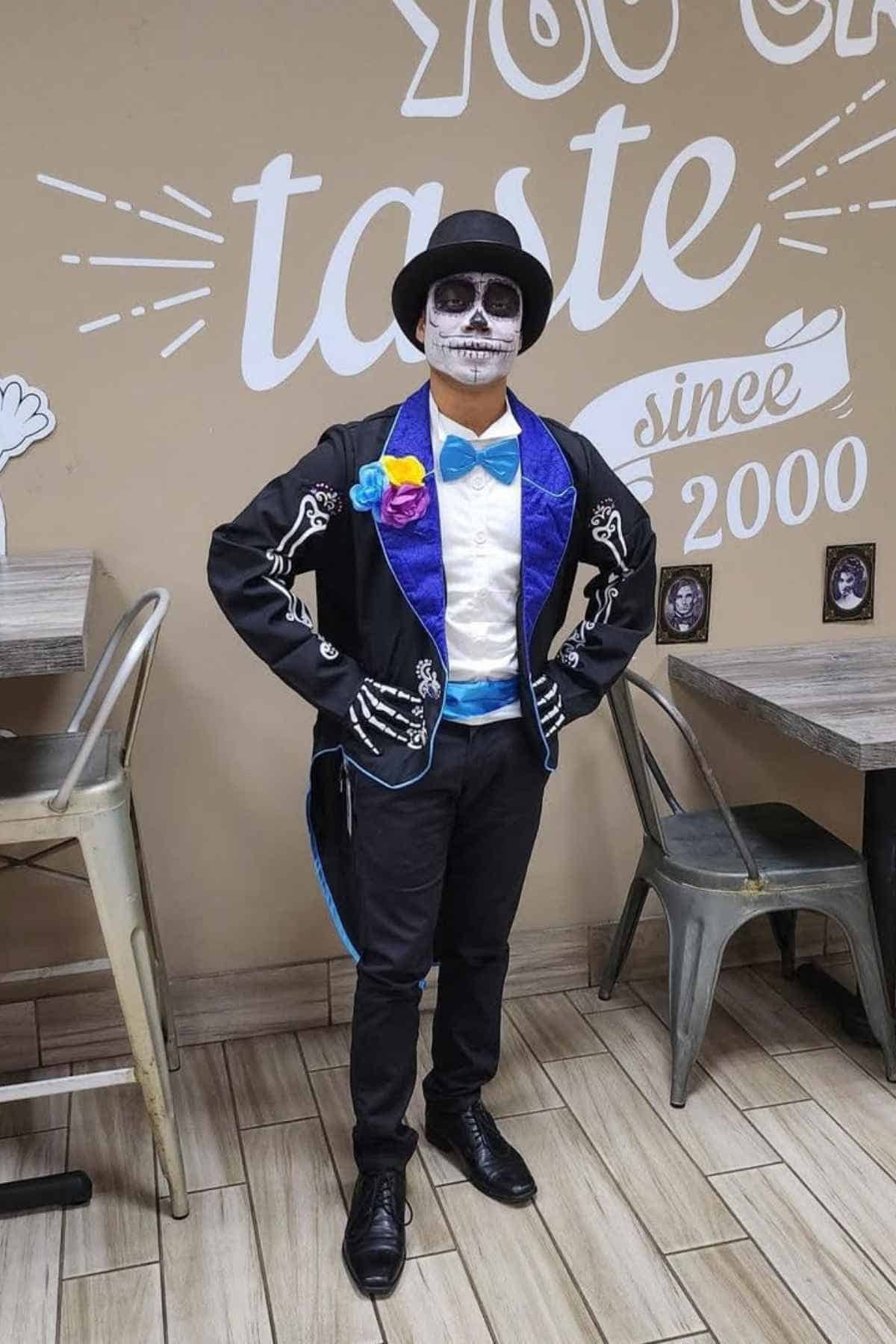 Person dressed in a Day of the Dead costume with face painted as a skull, wearing a black hat, black suit with skeleton designs, blue bow tie, and colorful flowers.