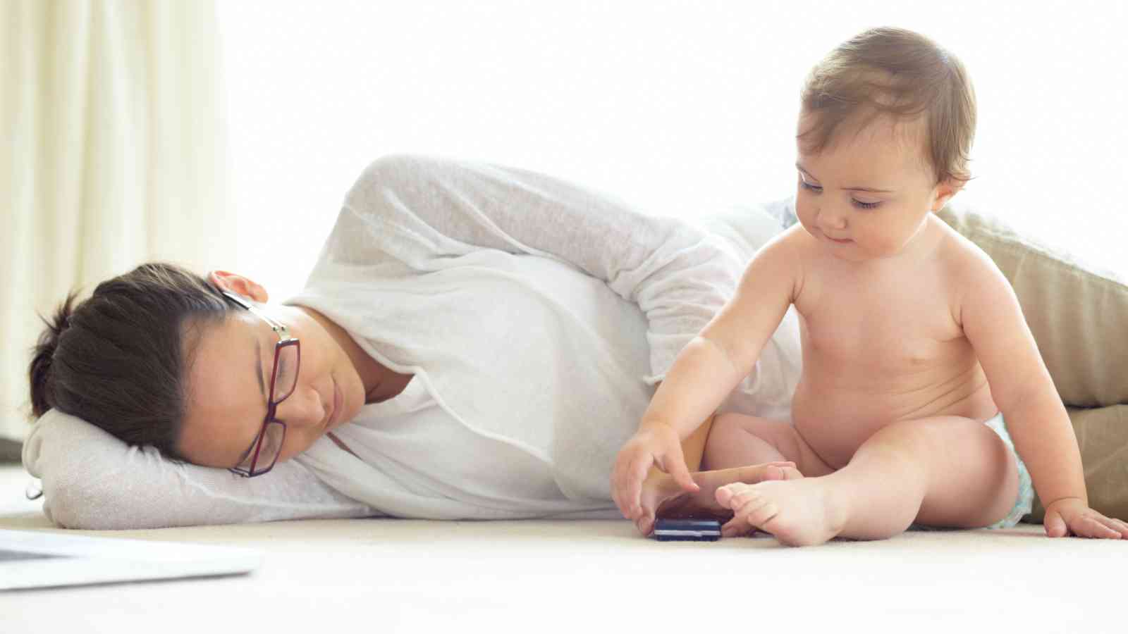An adult lies on the floor resting with eyes closed while a baby sitting nearby plays with a smartphone.