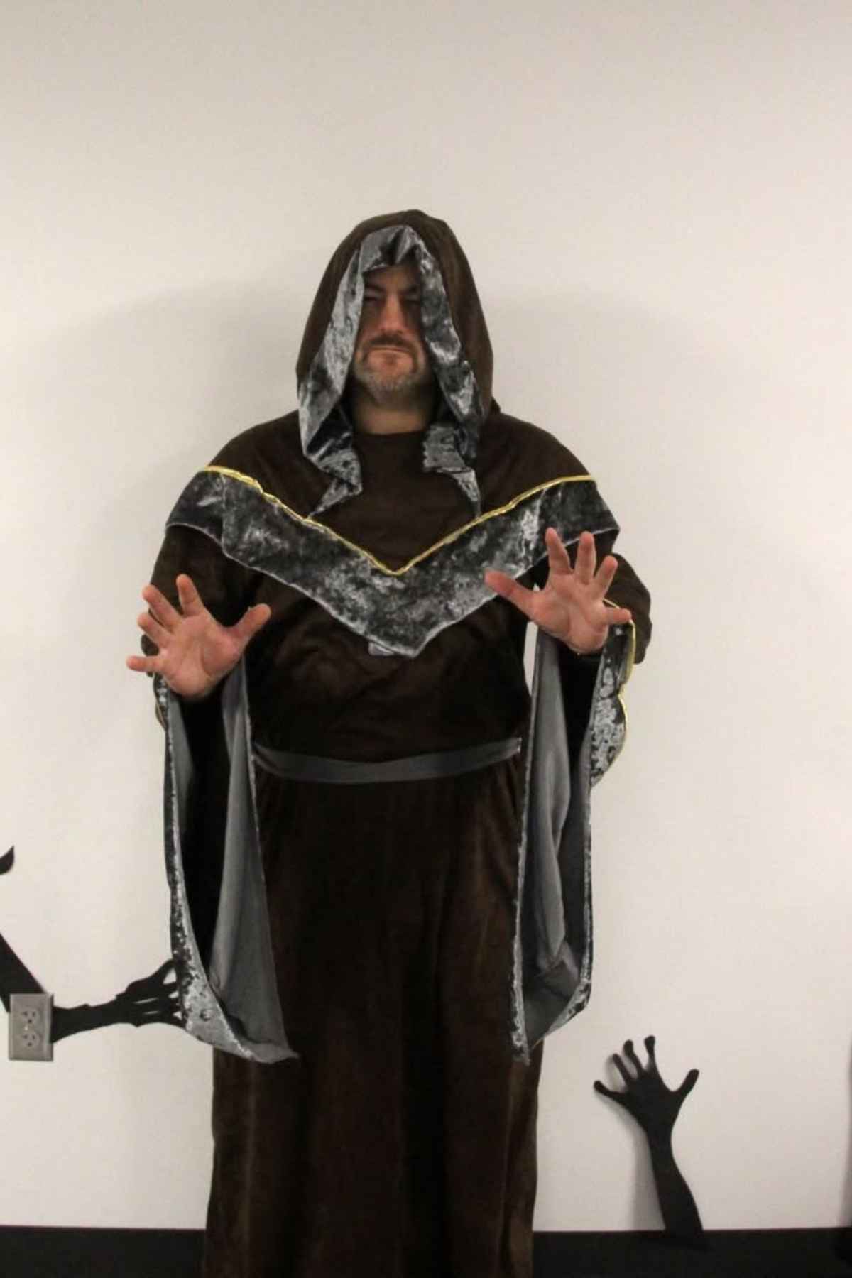 A person wearing a hooded brown robe with silver trim stands against a white wall, hands raised with palms forward. Black hand silhouettes are visible on the wall near the floor.