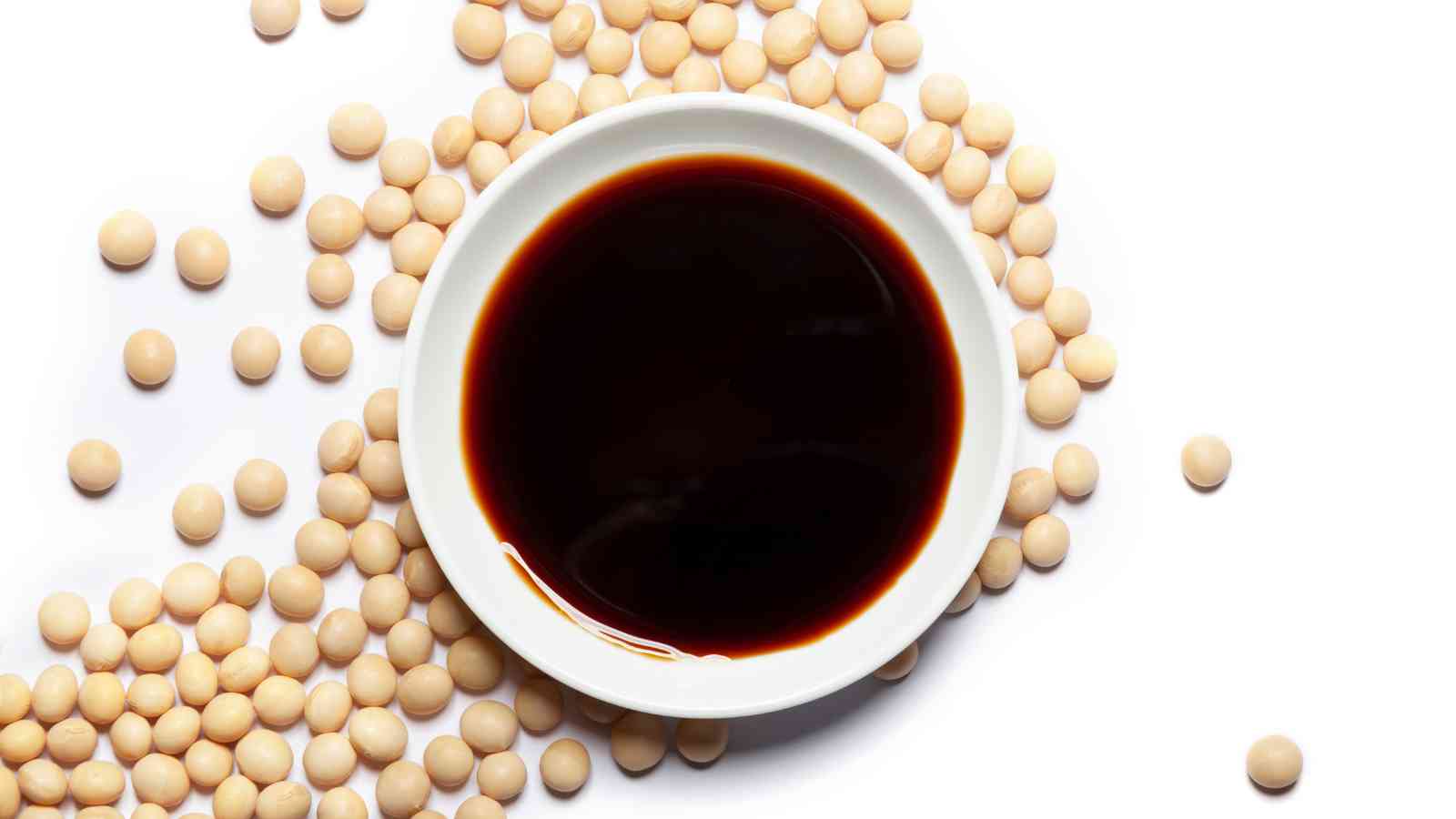 A white bowl filled with dark soy sauce is surrounded by raw soybeans scattered on a white surface.