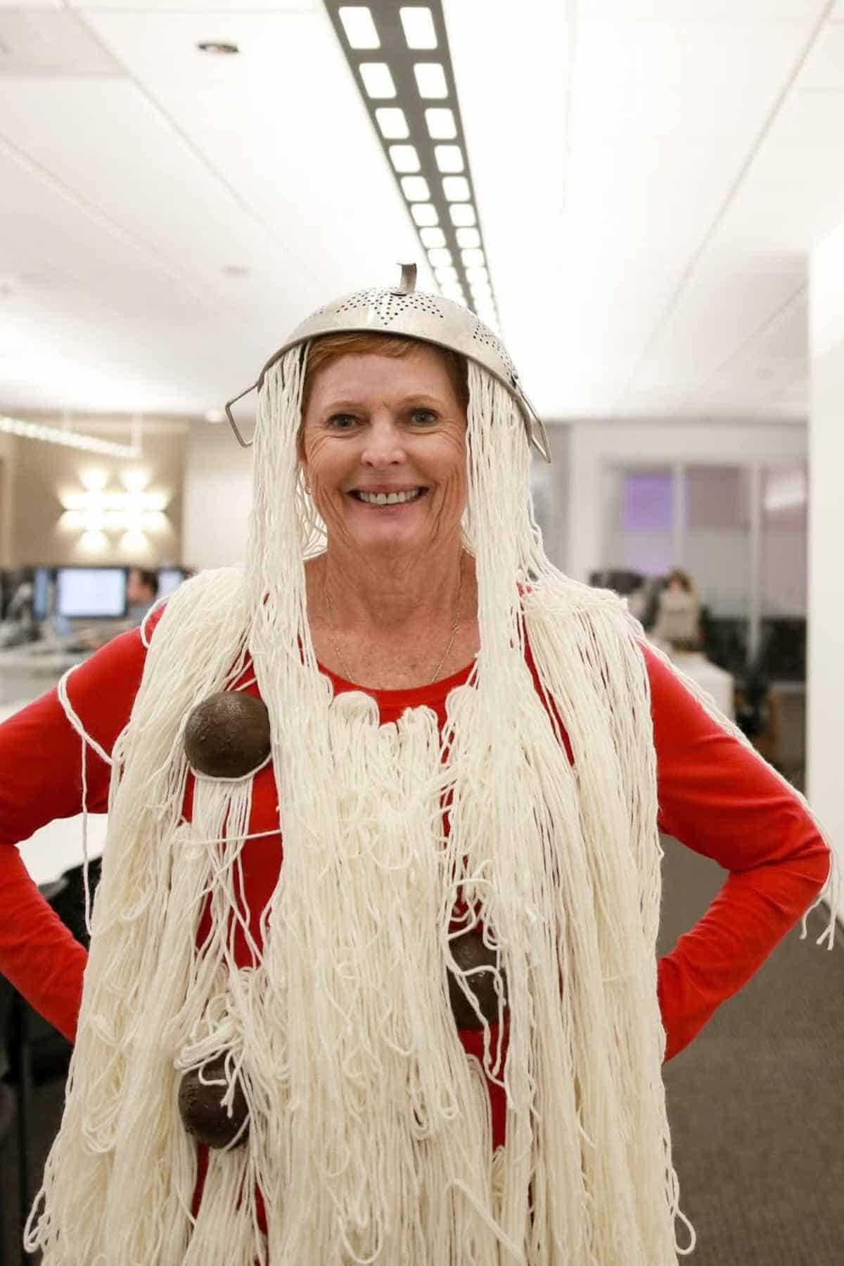 A woman in an office wears a red shirt with strands of white yarn draped over her shoulders, a metal colander on her head, and round objects attached to the yarn.