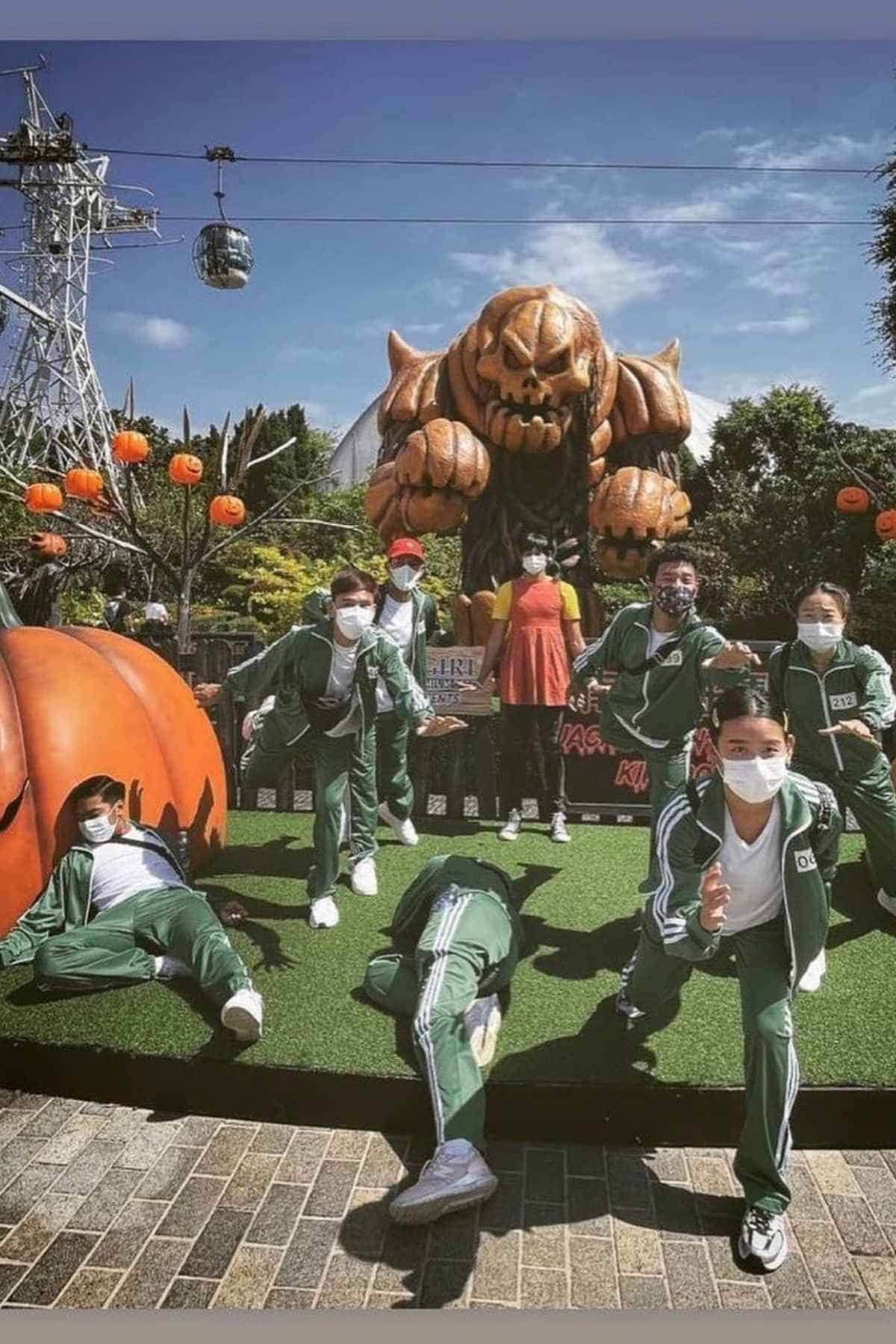 A group of teens in green tracksuits and face masks pose in front of a large monster statue and Halloween decorations at an outdoor amusement park.