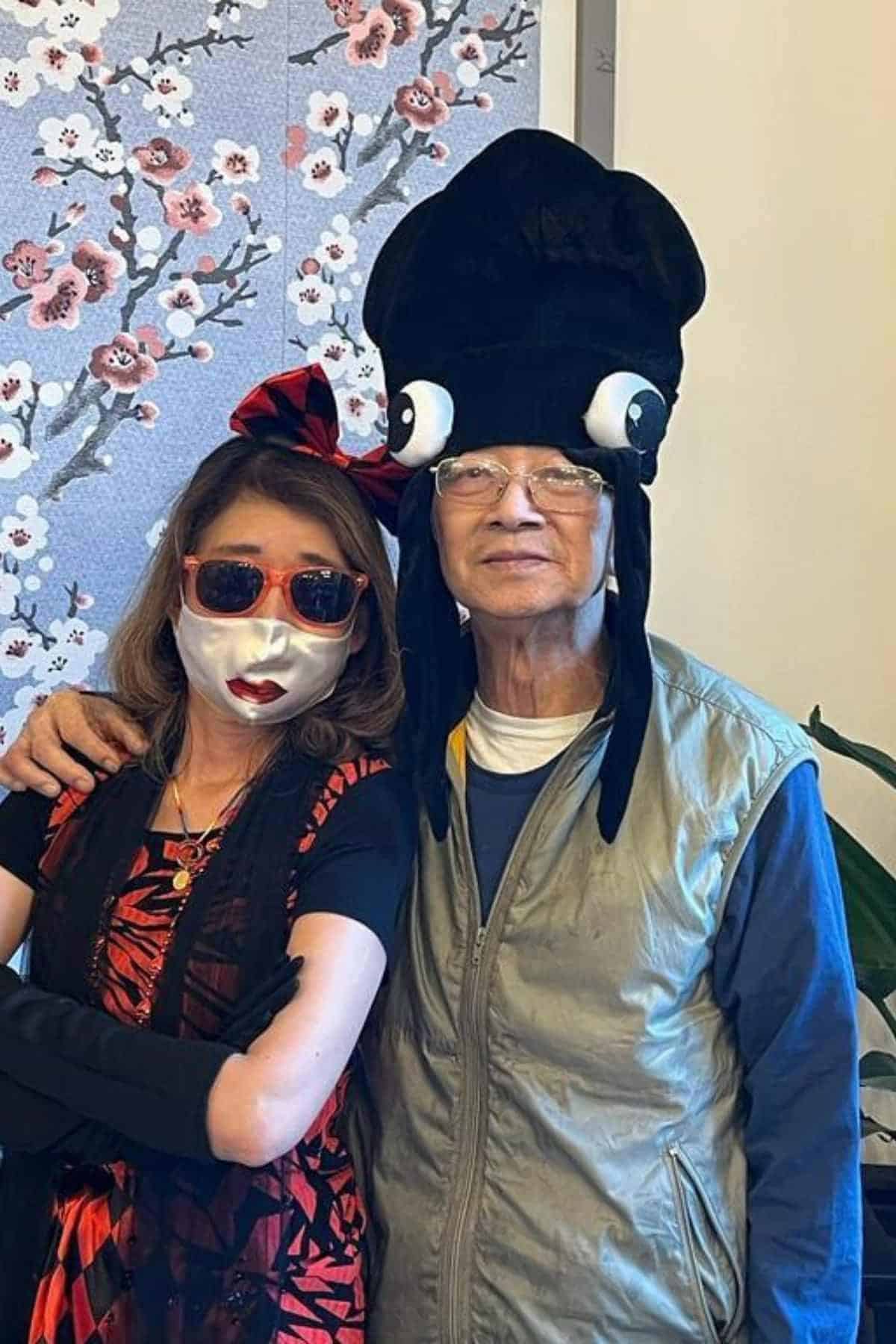 Two people pose together; one wears a white face mask, red sunglasses, and a red bow, while the other wears glasses and a large black squid-shaped hat.