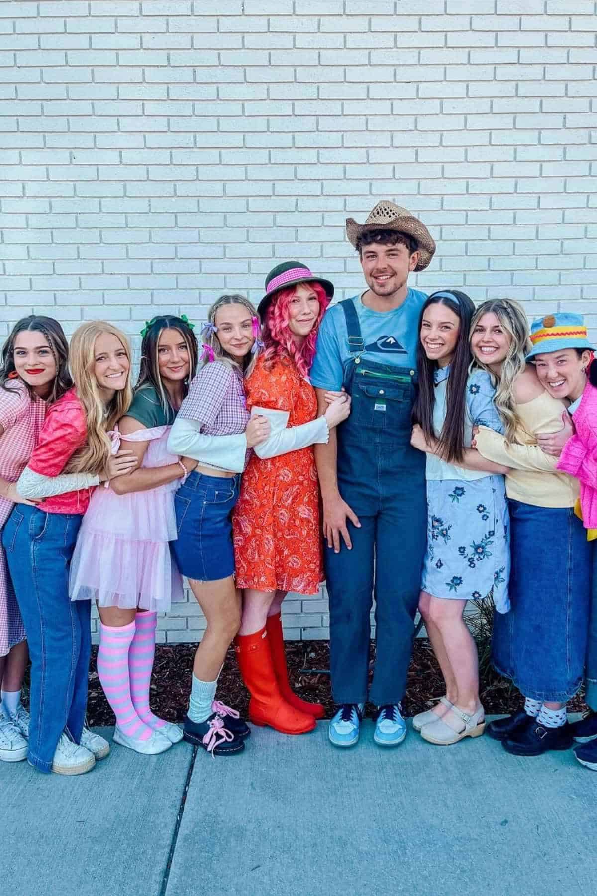 A group of nine people in colorful, vintage-style outfits stand in a row, smiling and hugging each other in front of a white brick wall.