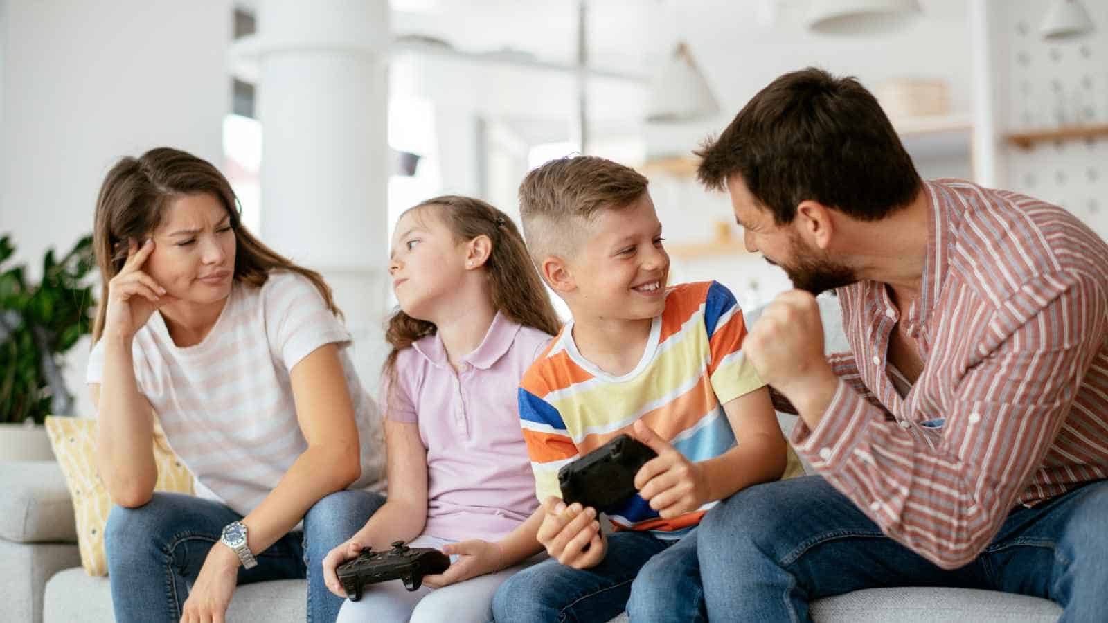 A family of four sits on a couch; two children hold video game controllers, the man appears excited, and the woman looks bored or annoyed.
