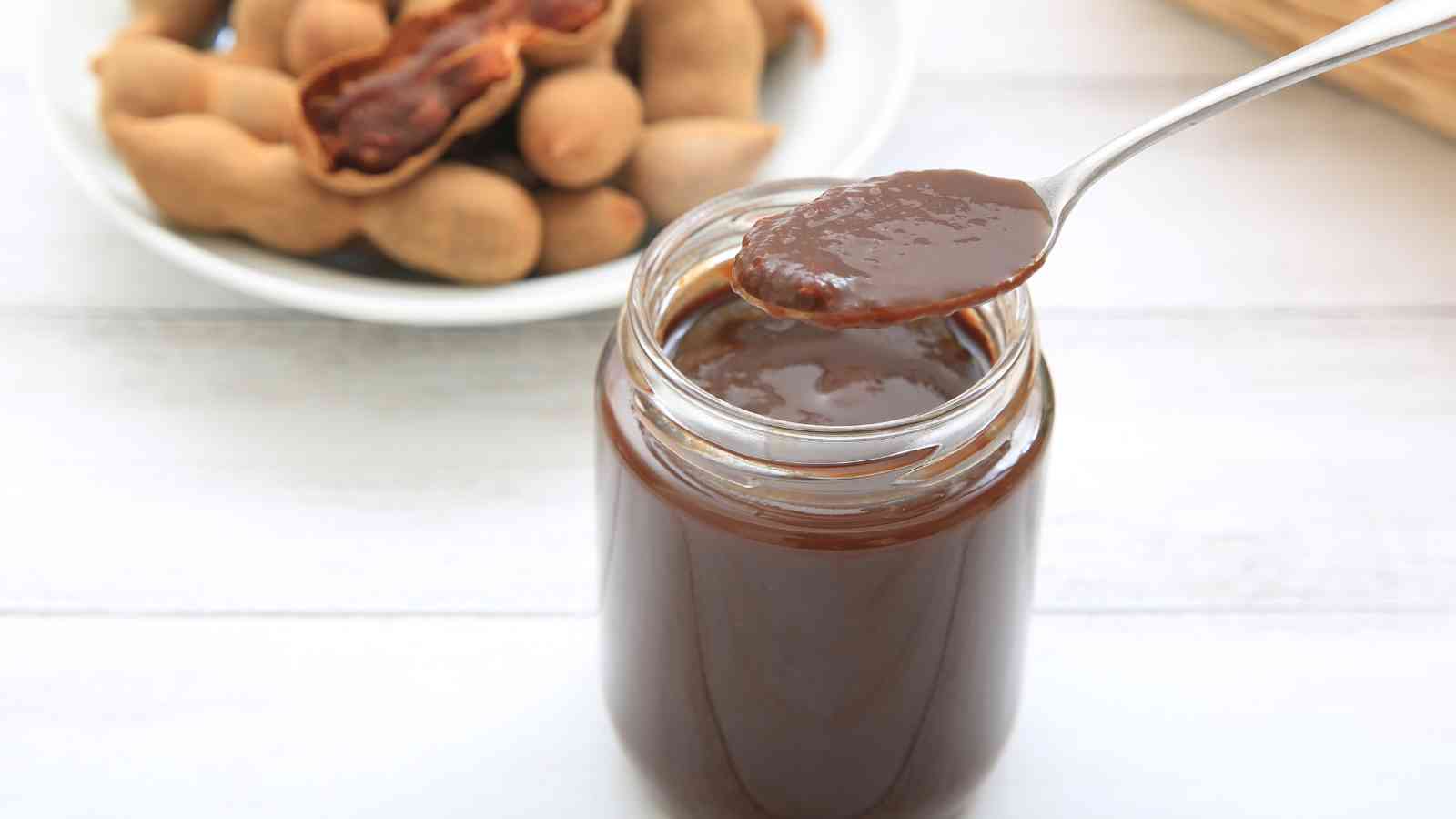 A jar of tamarind paste with a spoonful above it, and whole tamarind pods in a bowl in the background.
