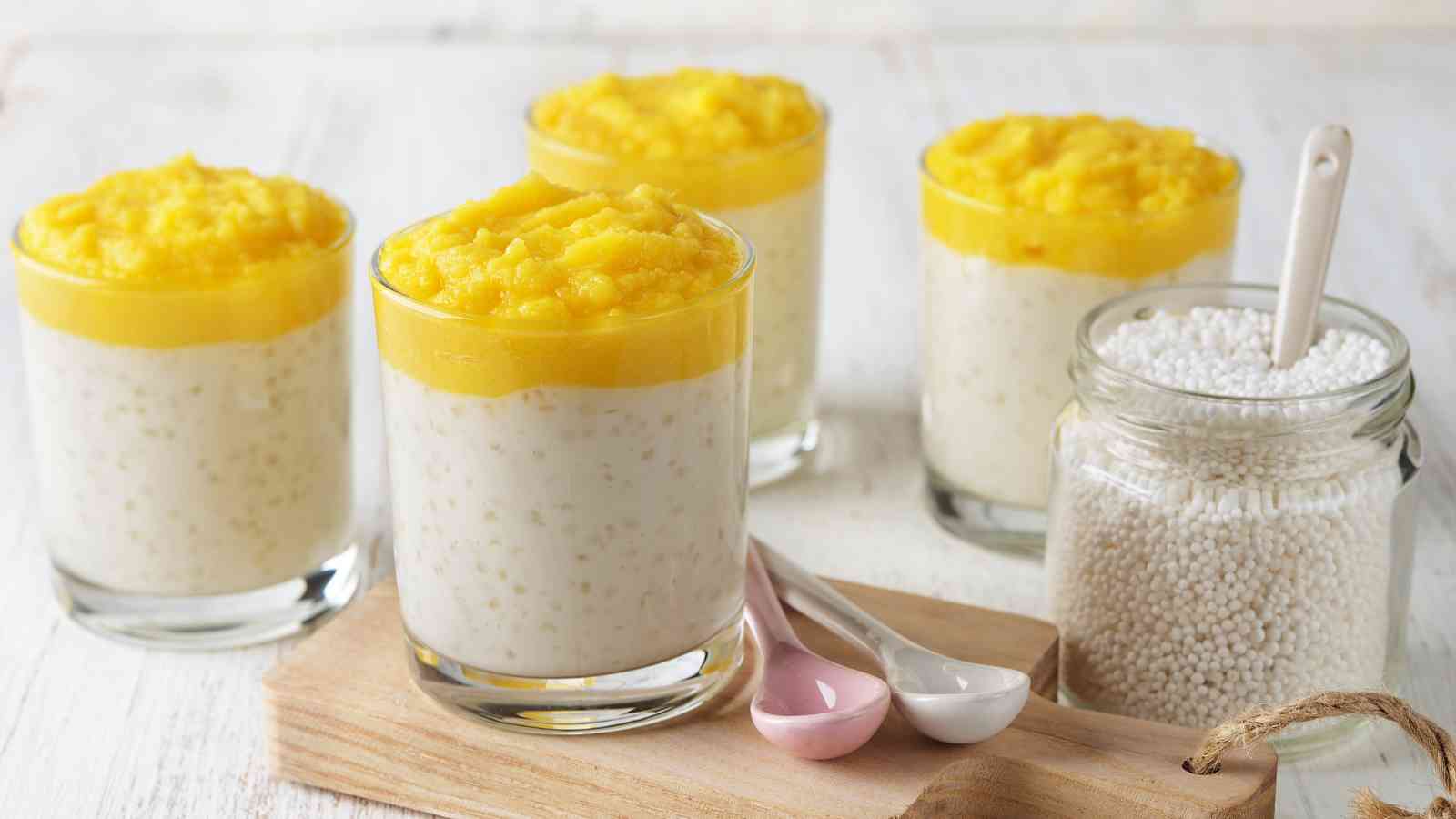 Five glasses of tapioca pudding topped with mango puree are arranged on a table next to a jar of tapioca pearls and three ceramic spoons.