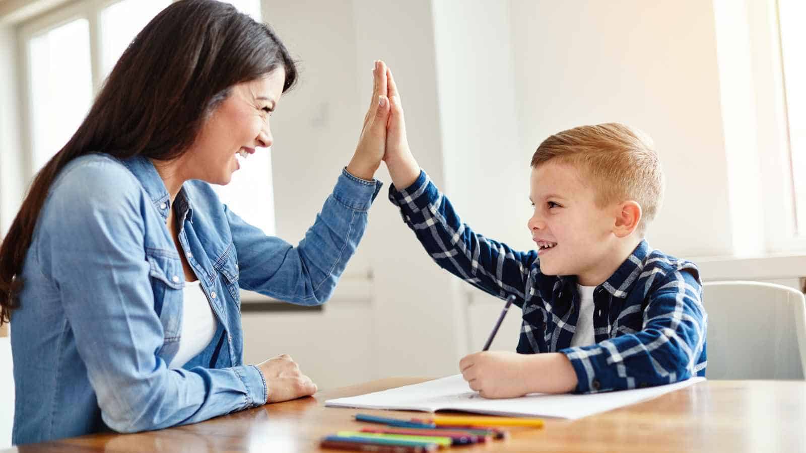 A woman and a young boy sitting at a table high-five each other, with colored pencils and paper in front of them.