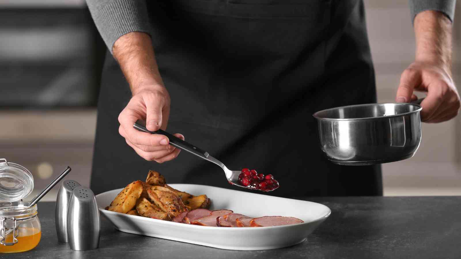 A person in a dark apron spooning cranberry sauce from a saucepan onto a plate with roasted potatoes and sliced meat.
