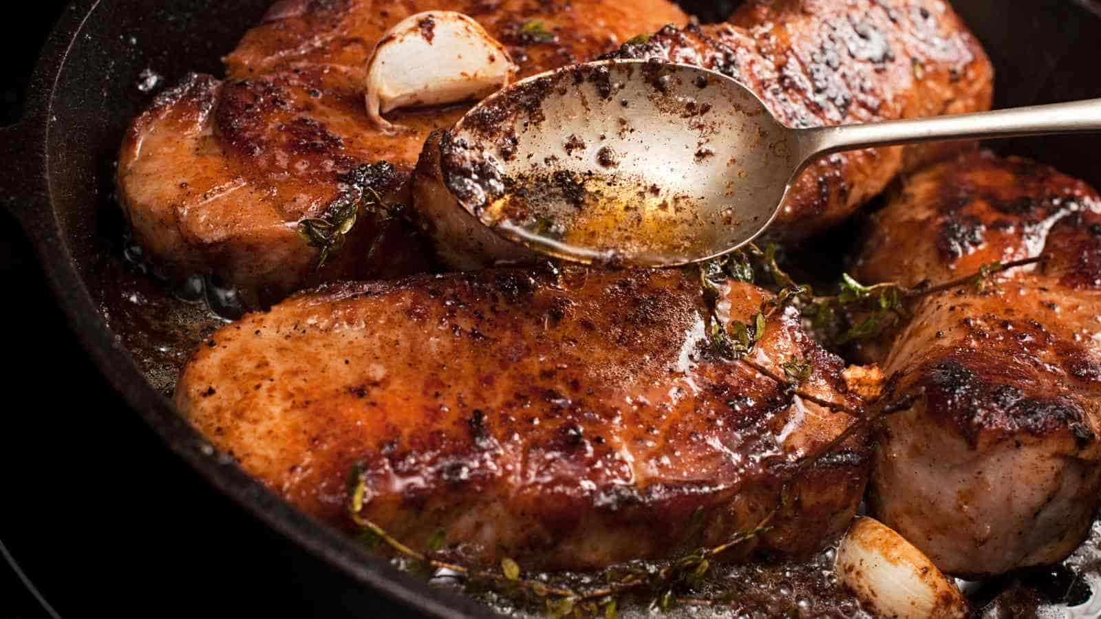 Several seasoned pork chops are being cooked in a skillet with garlic cloves, fresh herbs, and a spoon basting them with pan juices.