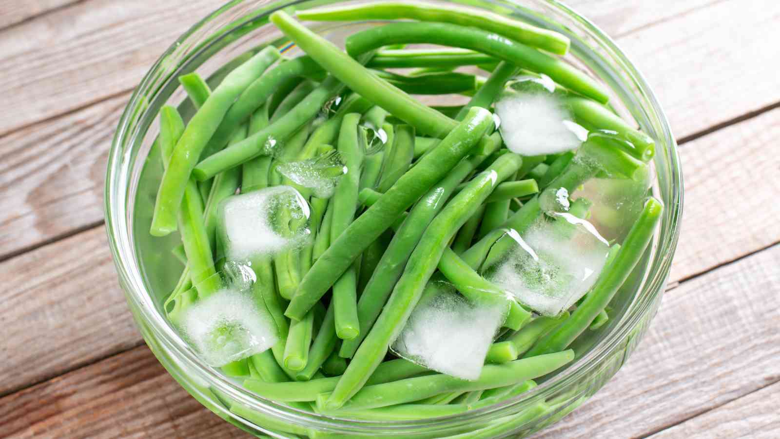 A glass bowl of green beans with ice cubes, sitting on a wooden surface.
