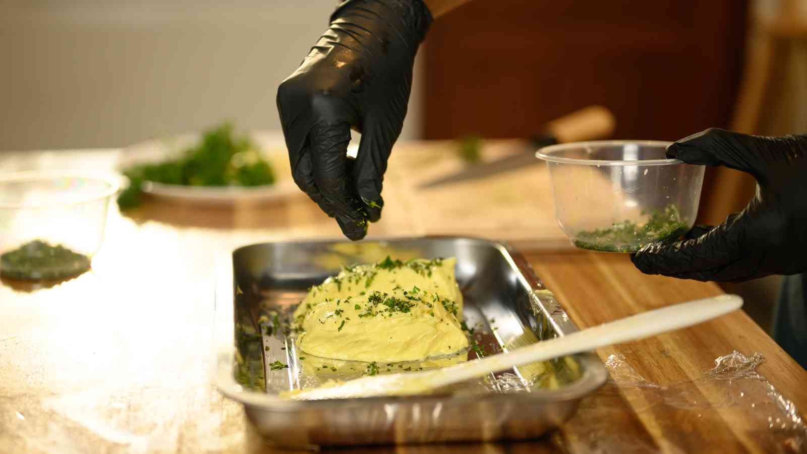 A person wearing black gloves sprinkles chopped herbs onto a mound of mashed potatoes in a metal tray on a wooden table.