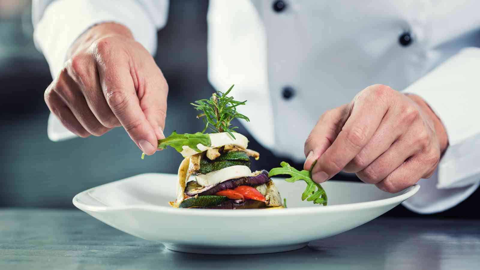 A chef’s hands are plating a dish with grilled vegetables and cheese, garnishing it with fresh greens on a white plate.