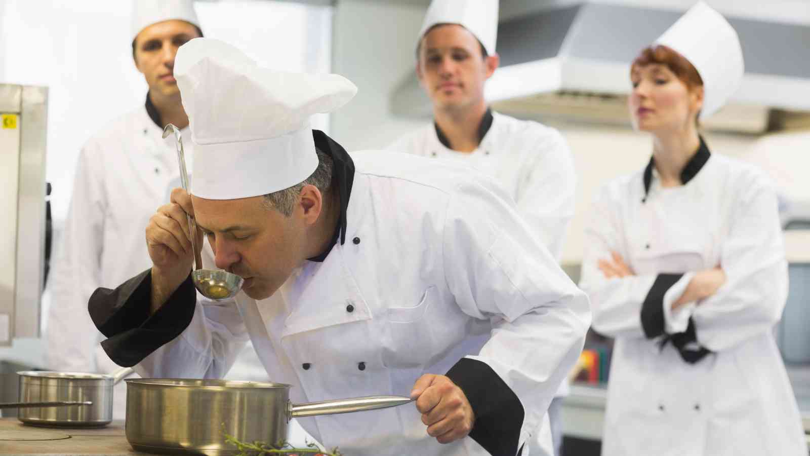 A chef in a white uniform and hat tastes food from a pot with a ladle, while three other chefs in similar uniforms watch attentively in a professional kitchen.