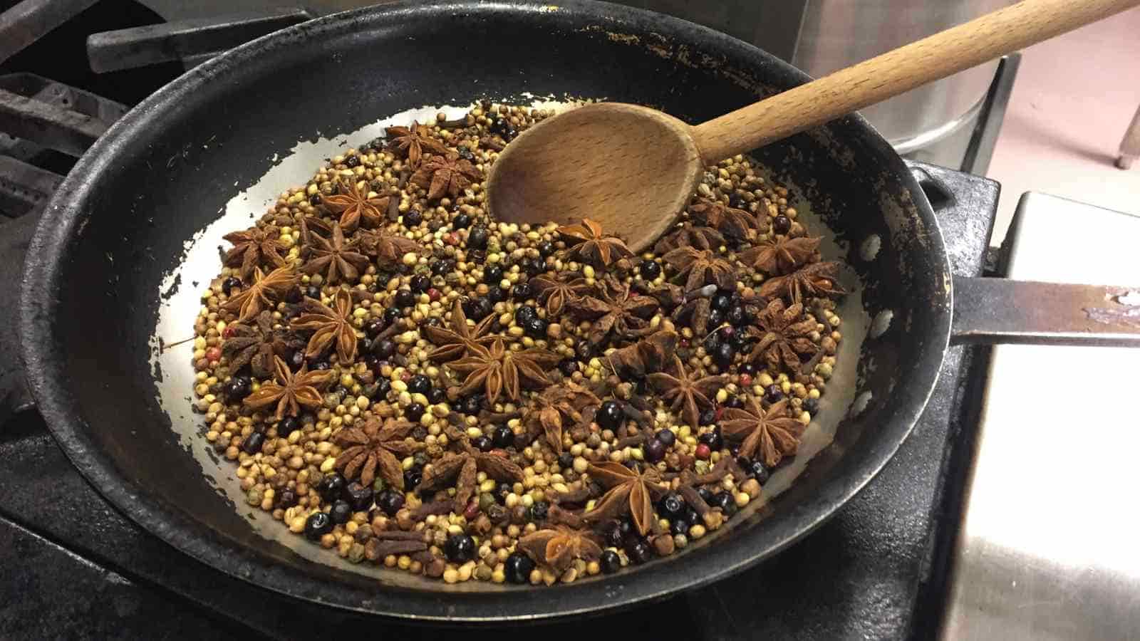 A black frying pan on a stove contains mixed whole spices, including star anise and coriander seeds, being stirred with a wooden spoon.