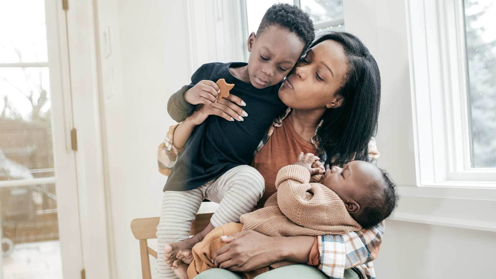 A woman sits by a window, holding a baby in her arms and kissing an older child on the cheek.