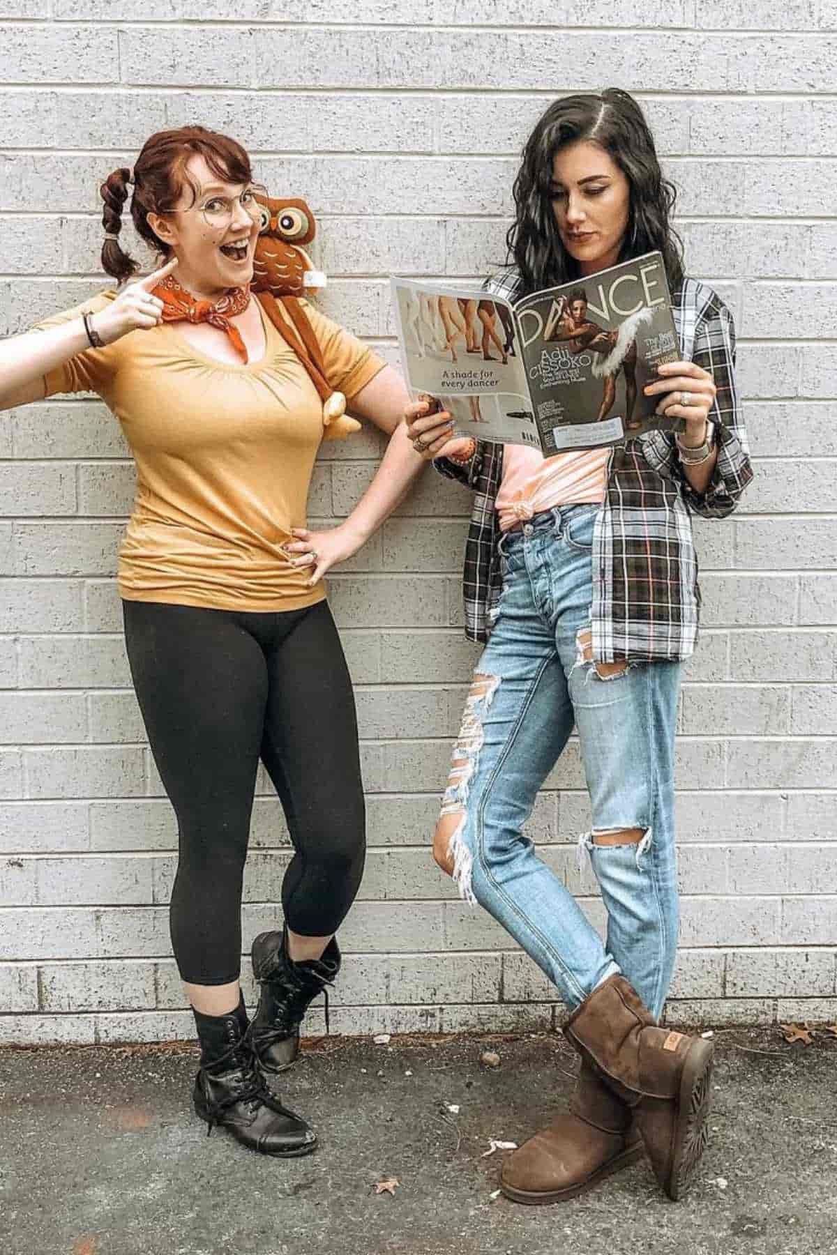 Two women stand against a white brick wall; one is dressed casually with a magazine, the other wears a yellow shirt, black leggings, and a toy monkey on her shoulder.