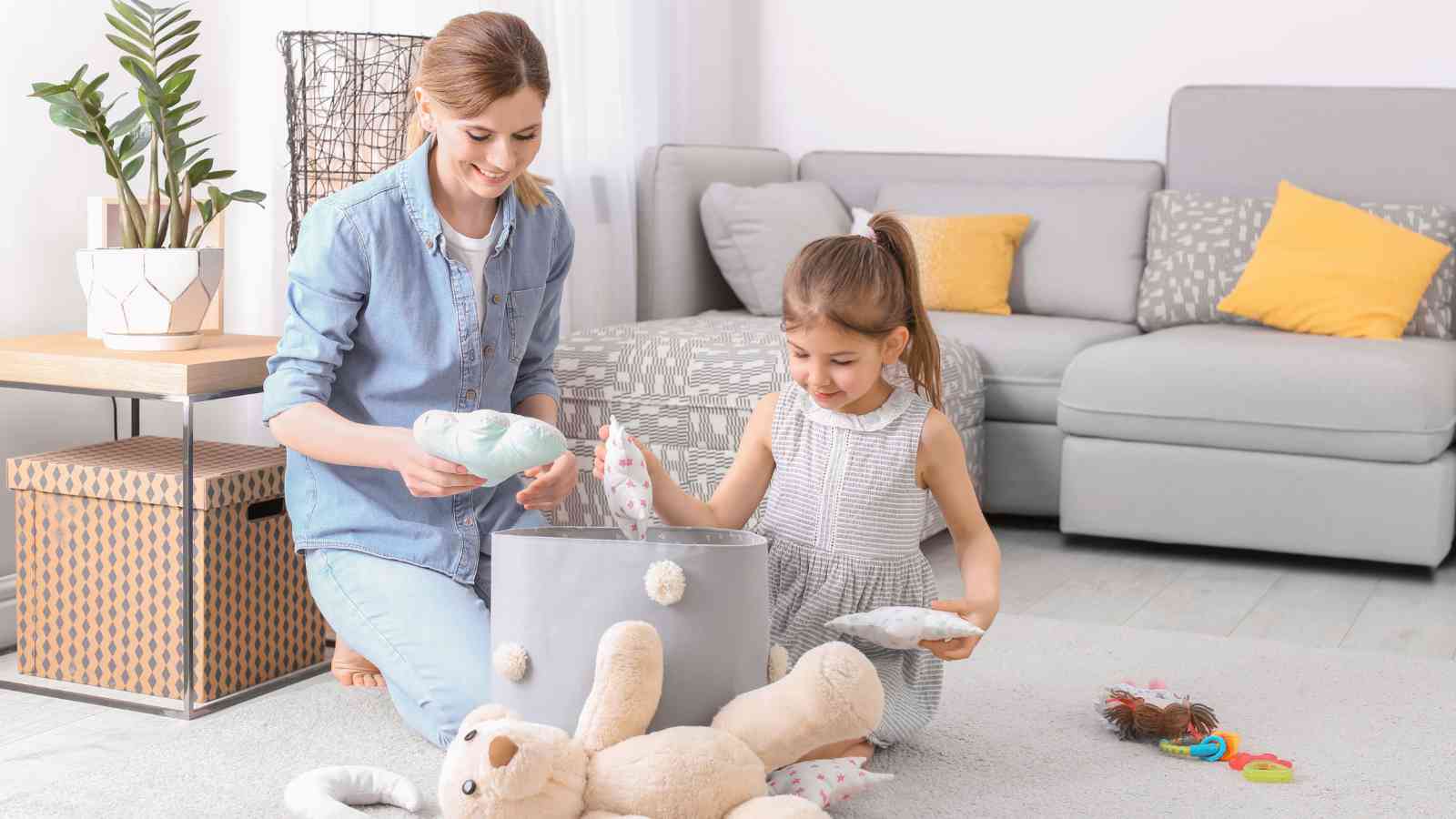 An adult and a child are kneeling on the floor in a living room, placing toys and stuffed animals into a storage bin.