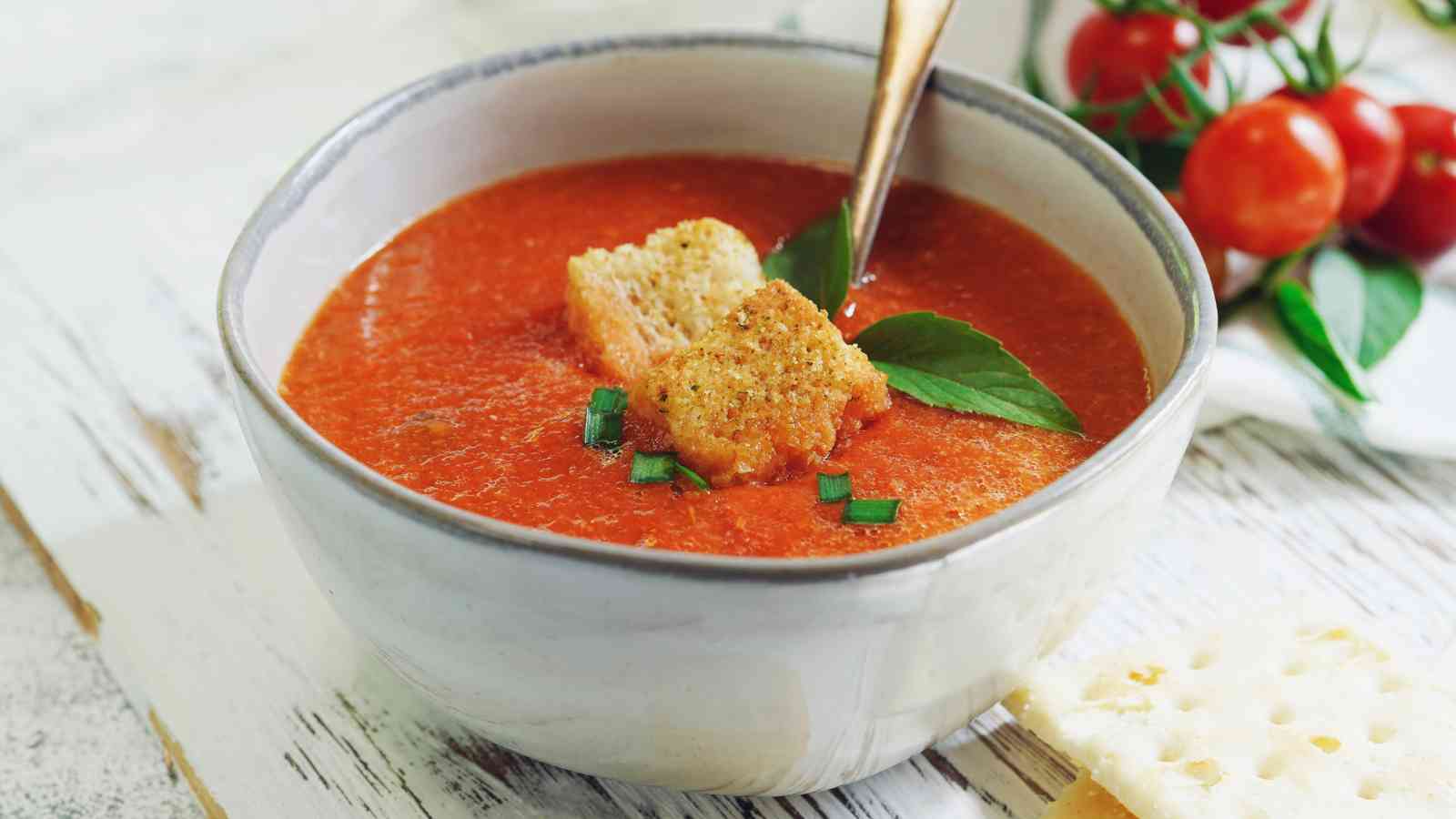 A bowl of tomato soup topped with croutons, chopped chives, and basil, with a spoon in the bowl and cherry tomatoes in the background.