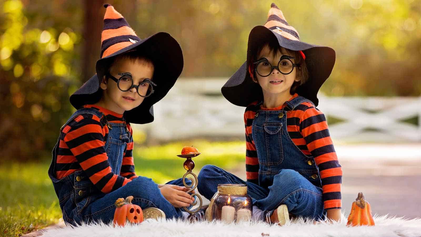 Two children in striped shirts, overalls, and witch hats sit outdoors on a blanket surrounded by Halloween decorations, including pumpkins and a jar—perfect inspiration for Halloween costumes for twin boys.