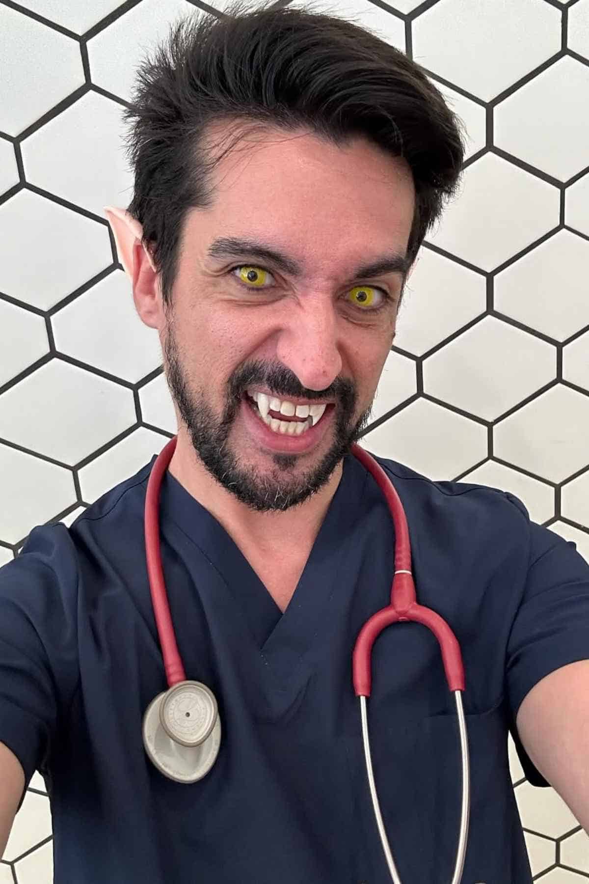 Person in medical scrubs with a stethoscope, showing fangs, yellow eyes, and pointed ears, standing in front of a hexagonal tile wall.