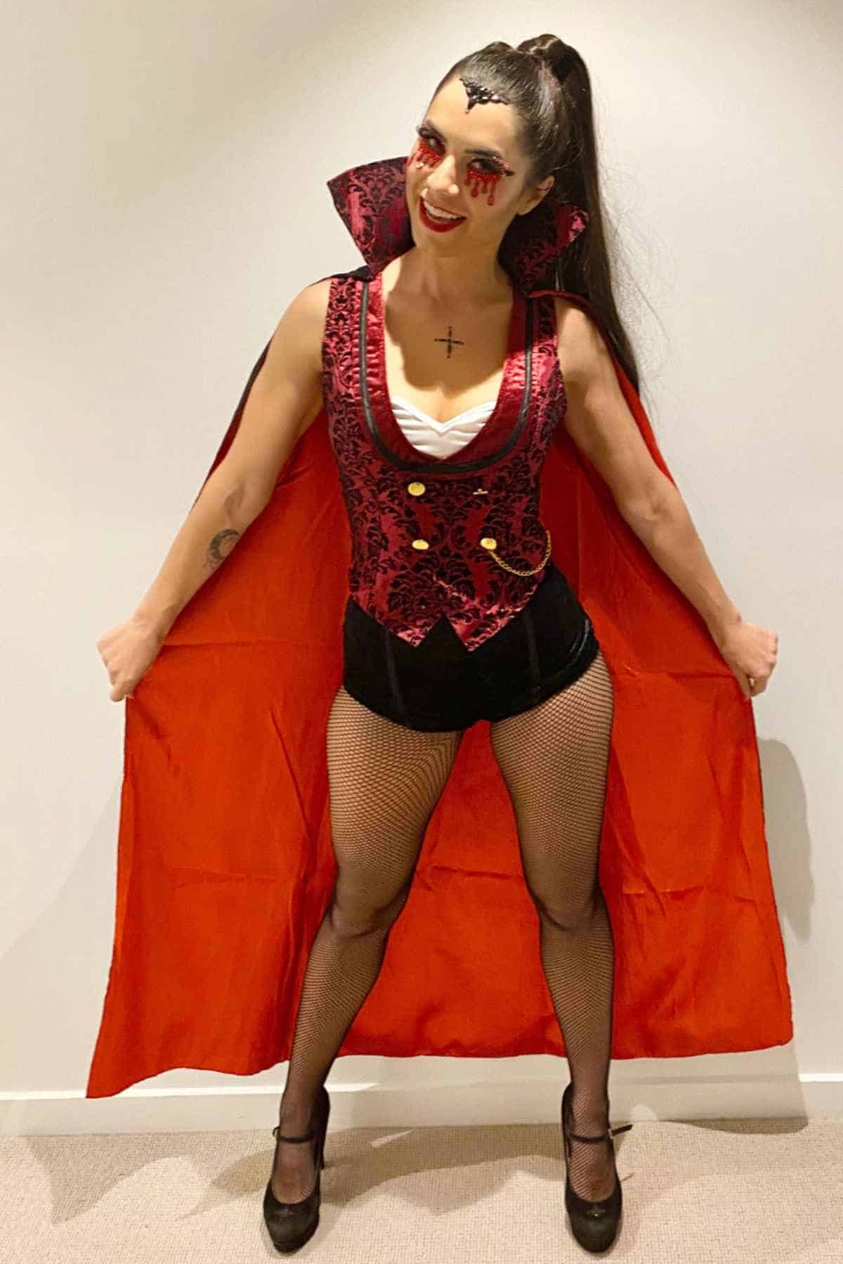 Person wearing a vampire costume with a red and black cape, fishnet stockings, dramatic makeup with fake blood, and black high heels, standing against a plain white wall.