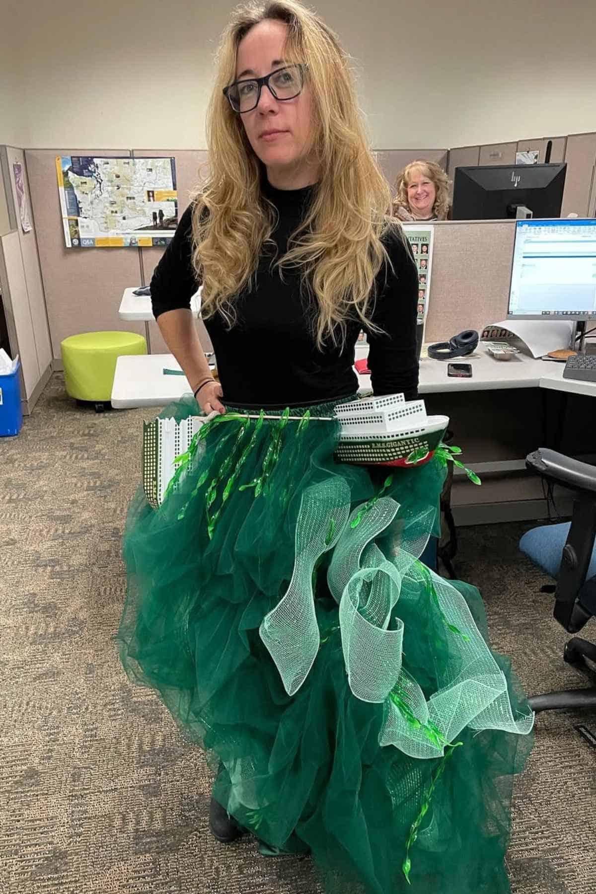 A woman in an office wears a costume resembling a green ocean wave with a model ship attached, while another person works at a desk in the background.