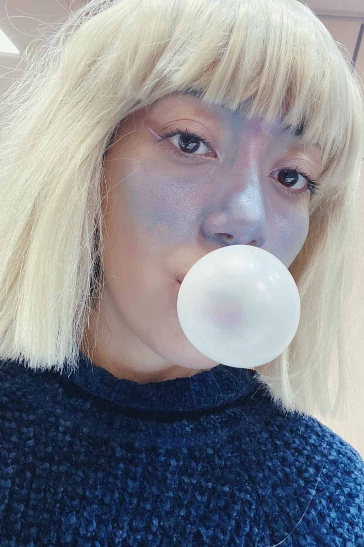 Person with blonde hair and blue face makeup blows a bubble with gum, wearing a dark textured sweater.