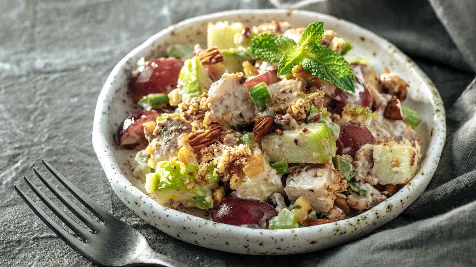 A plate of chicken salad with grapes, celery, apples, walnuts, and a creamy dressing, garnished with a mint sprig, next to a fork on a gray cloth.
