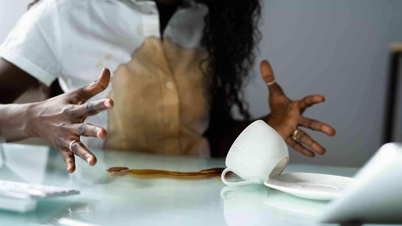 A person with coffee spilled on their shirt gestures in frustration, next to a tipped-over coffee cup and spilled coffee on a desk.