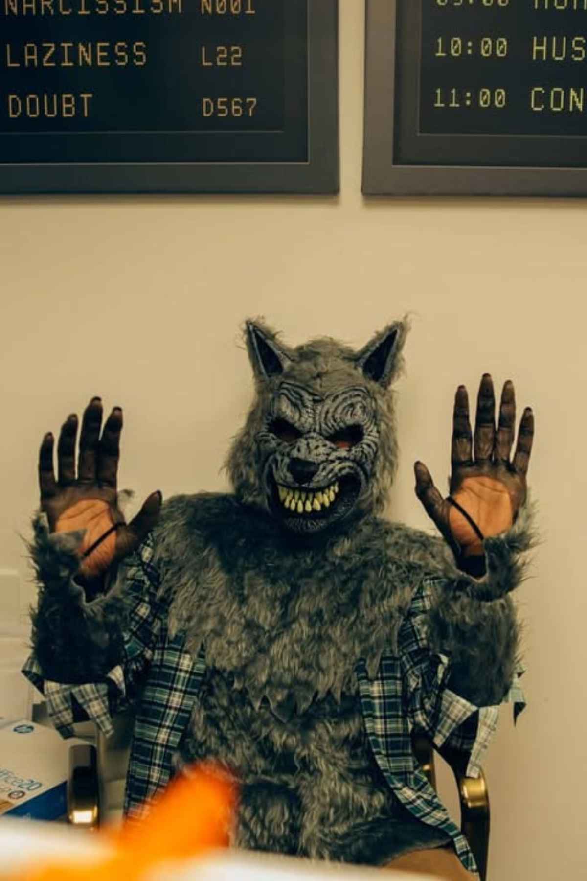 Person wearing a werewolf costume with raised hands sits indoors; office signs with text are visible on the wall behind them.