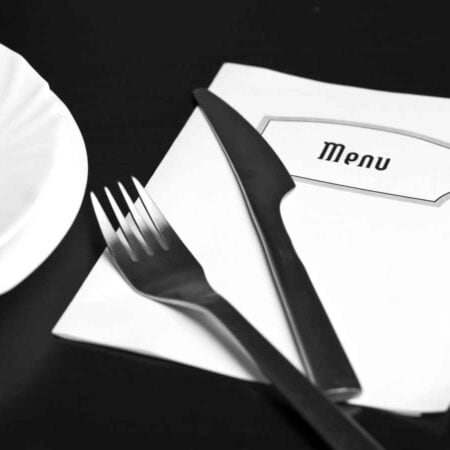 A white plate sits next to a fork, knife, and a folded menu on a dark surface.