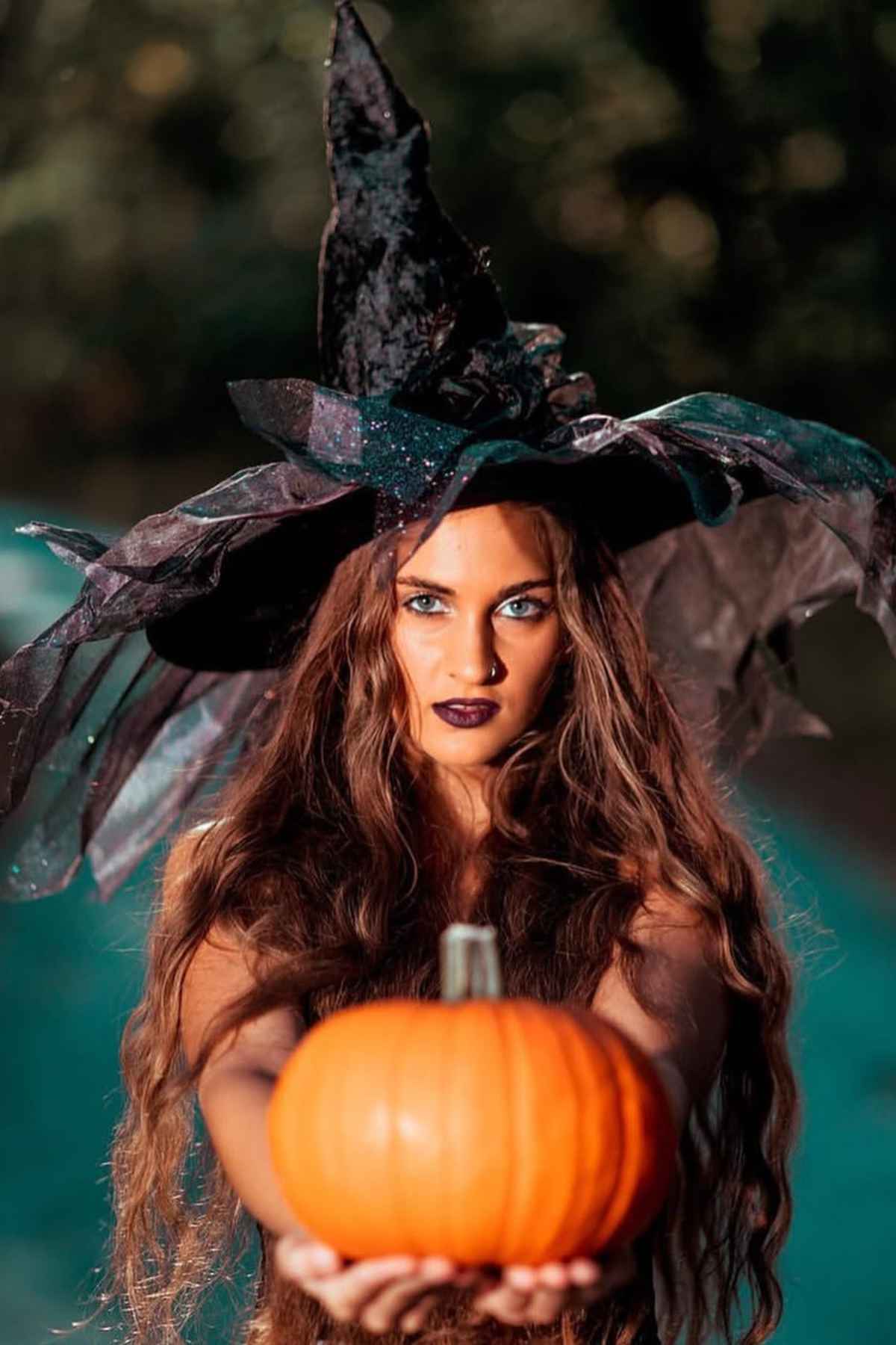 Woman in a black witch hat holds a small orange pumpkin towards the camera, with long wavy hair and dark makeup, outdoors.