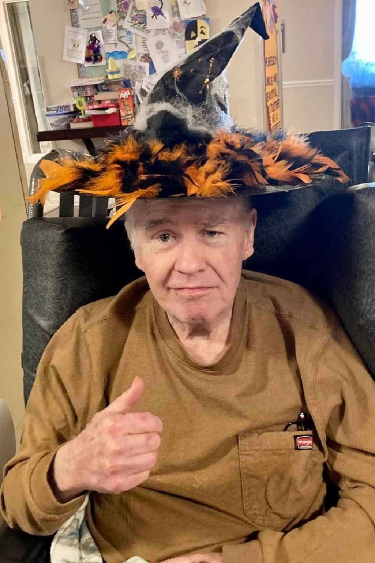 An older man sits in a chair, wearing a brown shirt and a festive witch hat with orange and black decorations, giving a thumbs-up. Cards and notes are visible on the wall behind him.