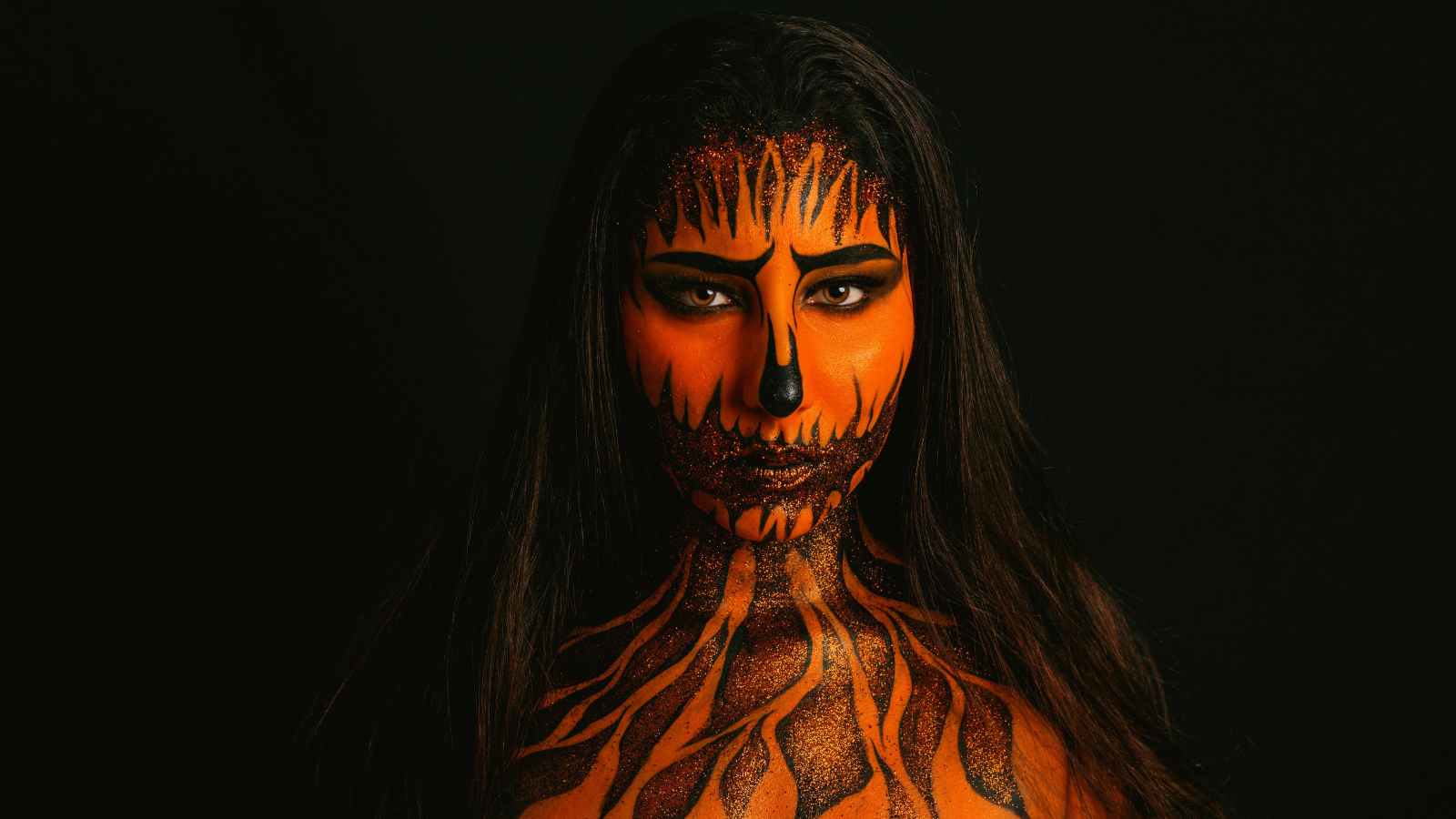 A person with long dark hair, painted with orange and black makeup resembling flames or tiger stripes, poses against a black background.