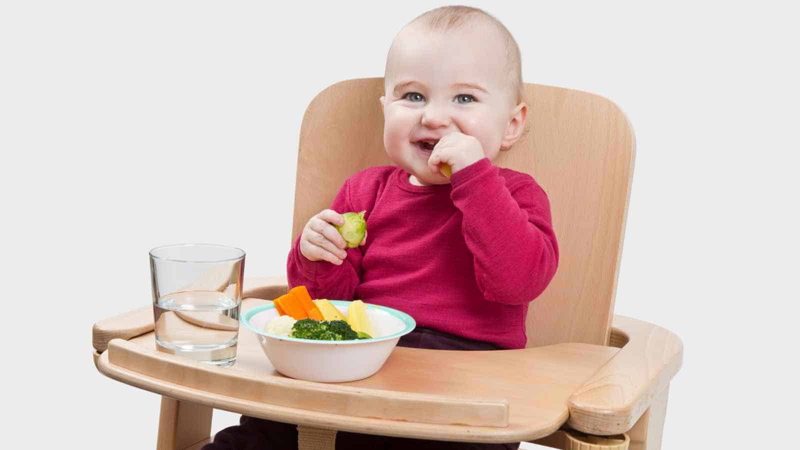 A baby in a red shirt sits in a wooden high chair, eating vegetables from a bowl with a glass of water on the tray.