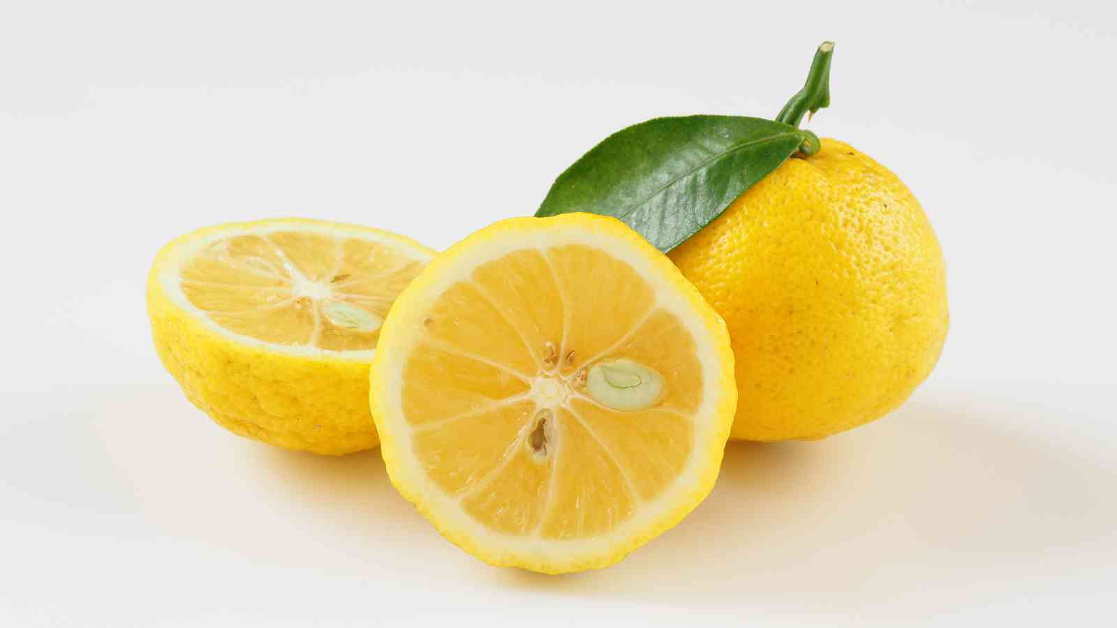 A whole yuzu fruit with a green leaf and two halves of yuzu, showing pale yellow flesh and seeds, set against a white background.