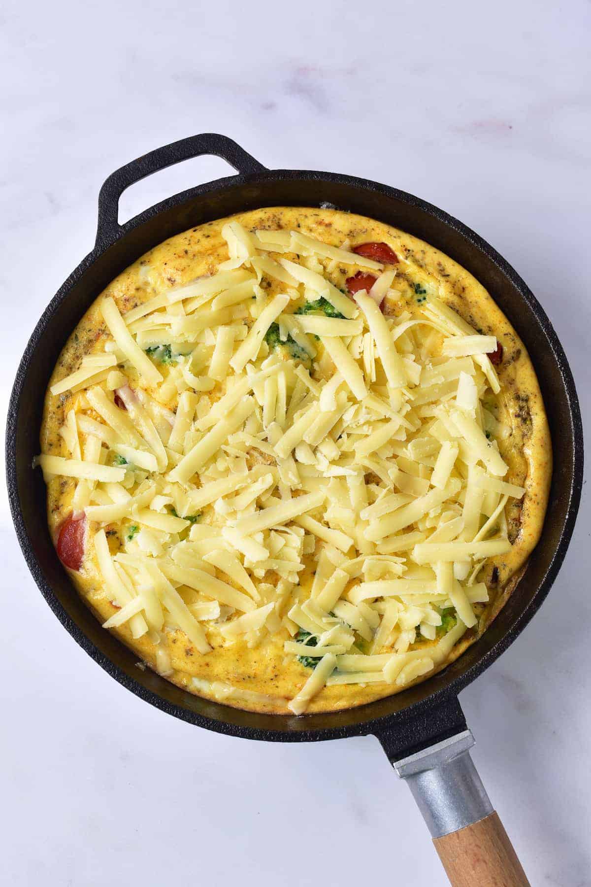 A black skillet contains an uncooked frittata topped with shredded cheese, visible vegetables, and slices of sausage on a white surface.
