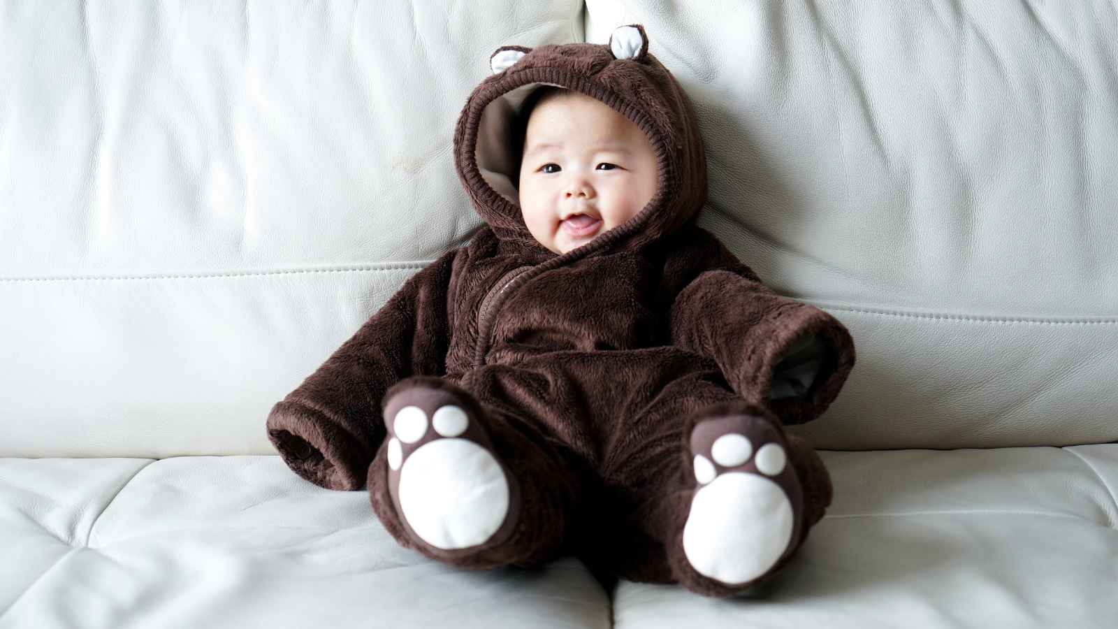 Baby wearing a fluffy bear suit while sitting in a couch.