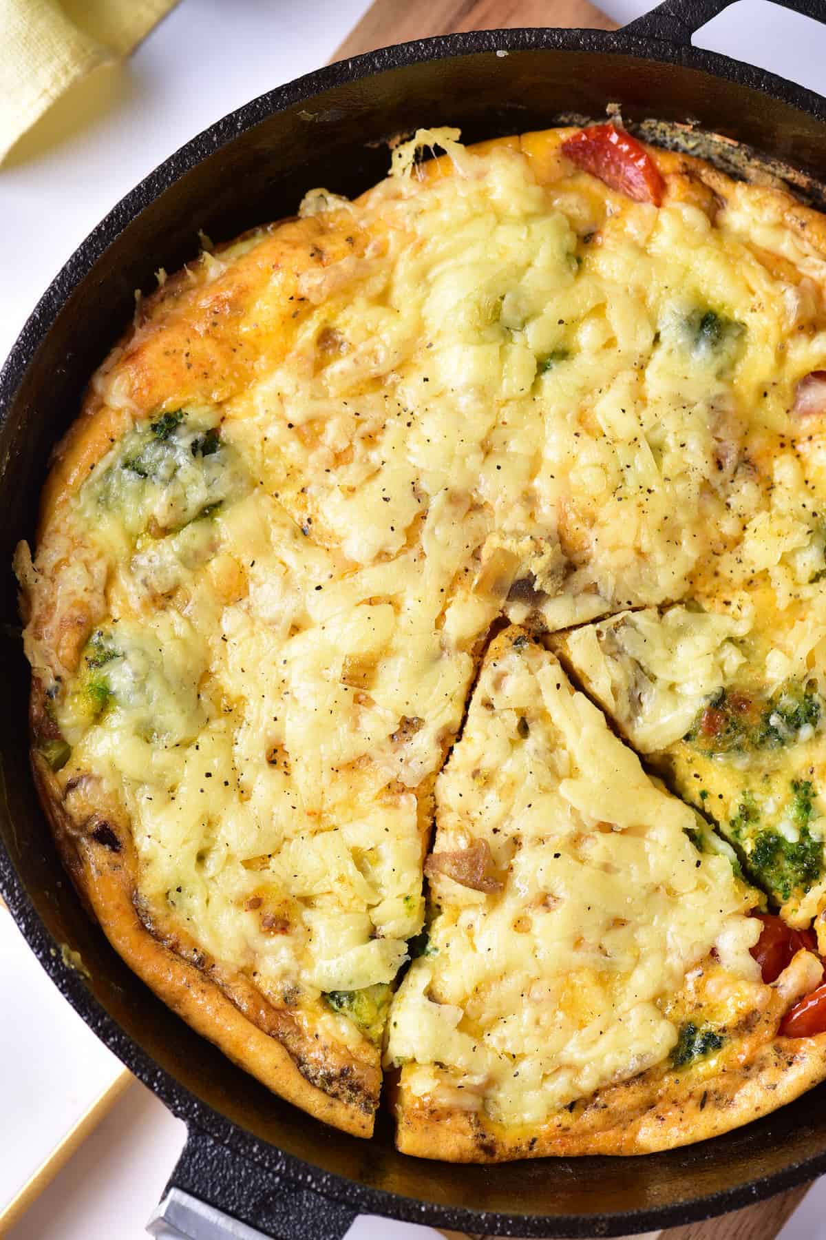A baked frittata in a skillet with melted cheese on top, containing visible vegetables and a slice cut out.