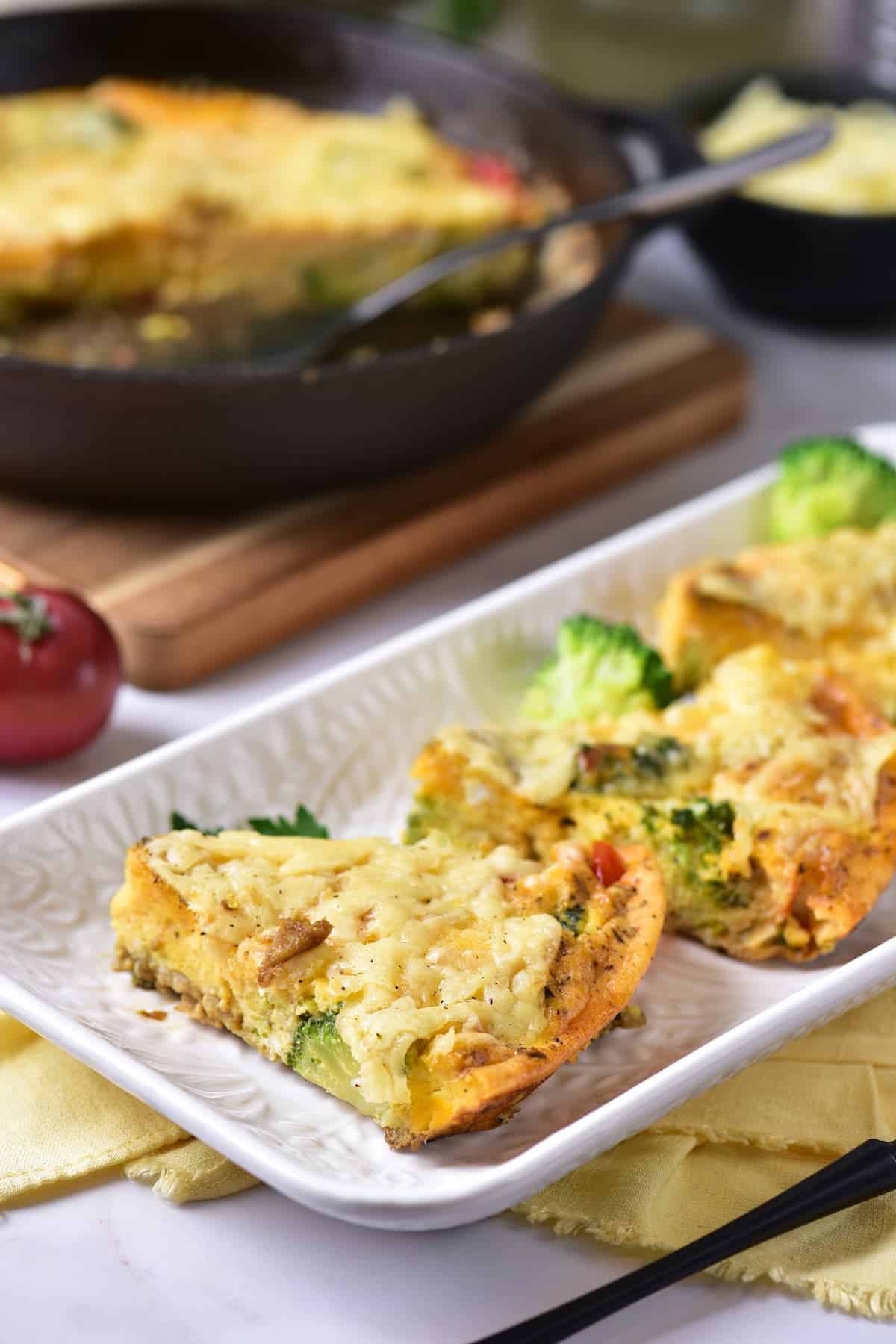 Two slices of vegetable frittata with cheese on a white rectangular plate, with more frittata in a skillet in the background.