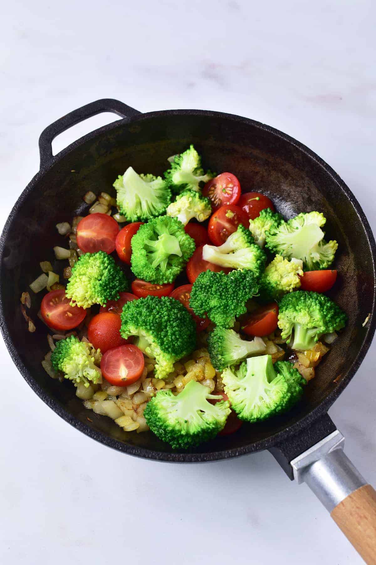 Broccoli florets, halved cherry tomatoes, and sautéed onions cooking in a black skillet on a white surface.