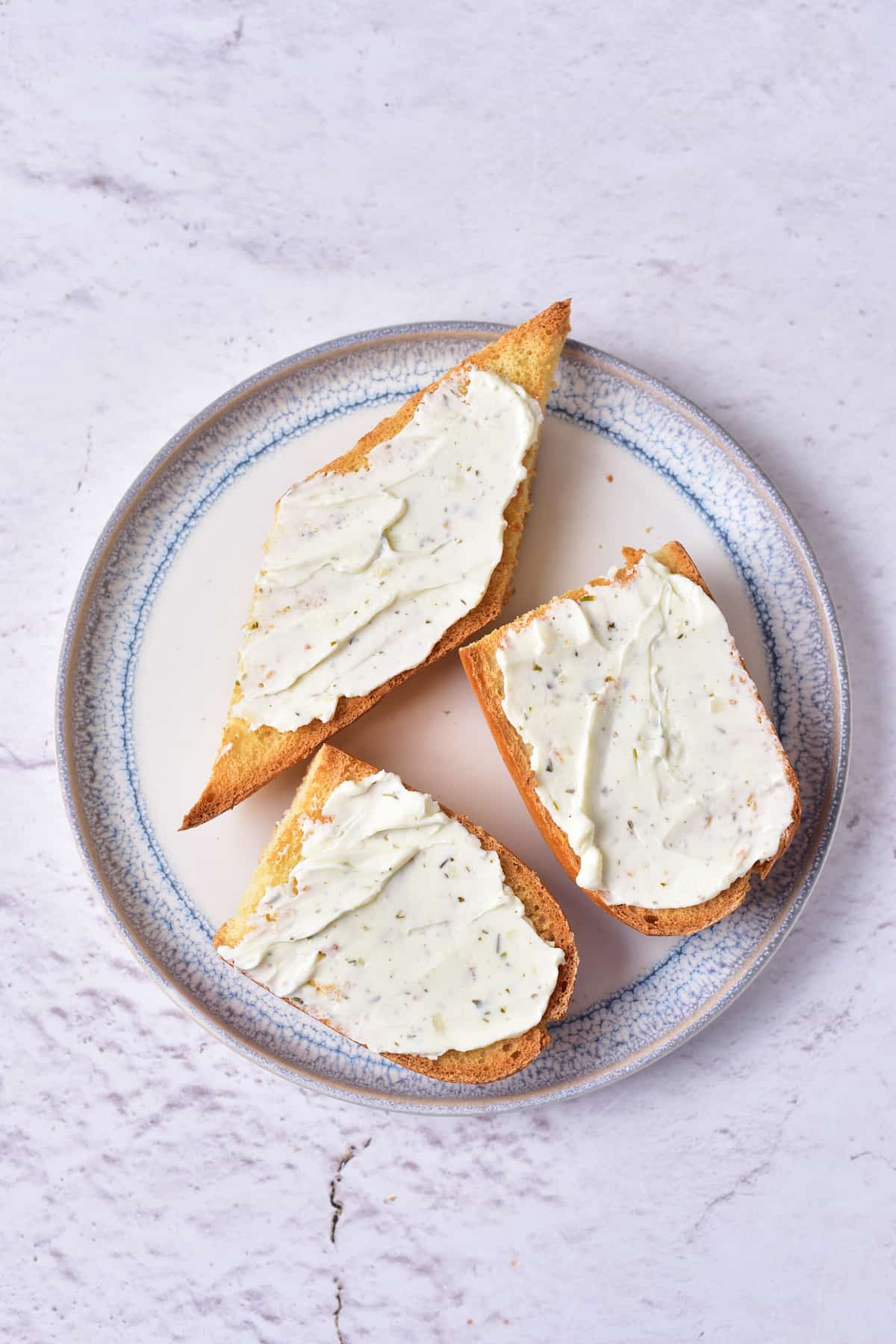 Three slices of toasted bread with a layer of herbed cream cheese on a round plate, set on a light textured surface.