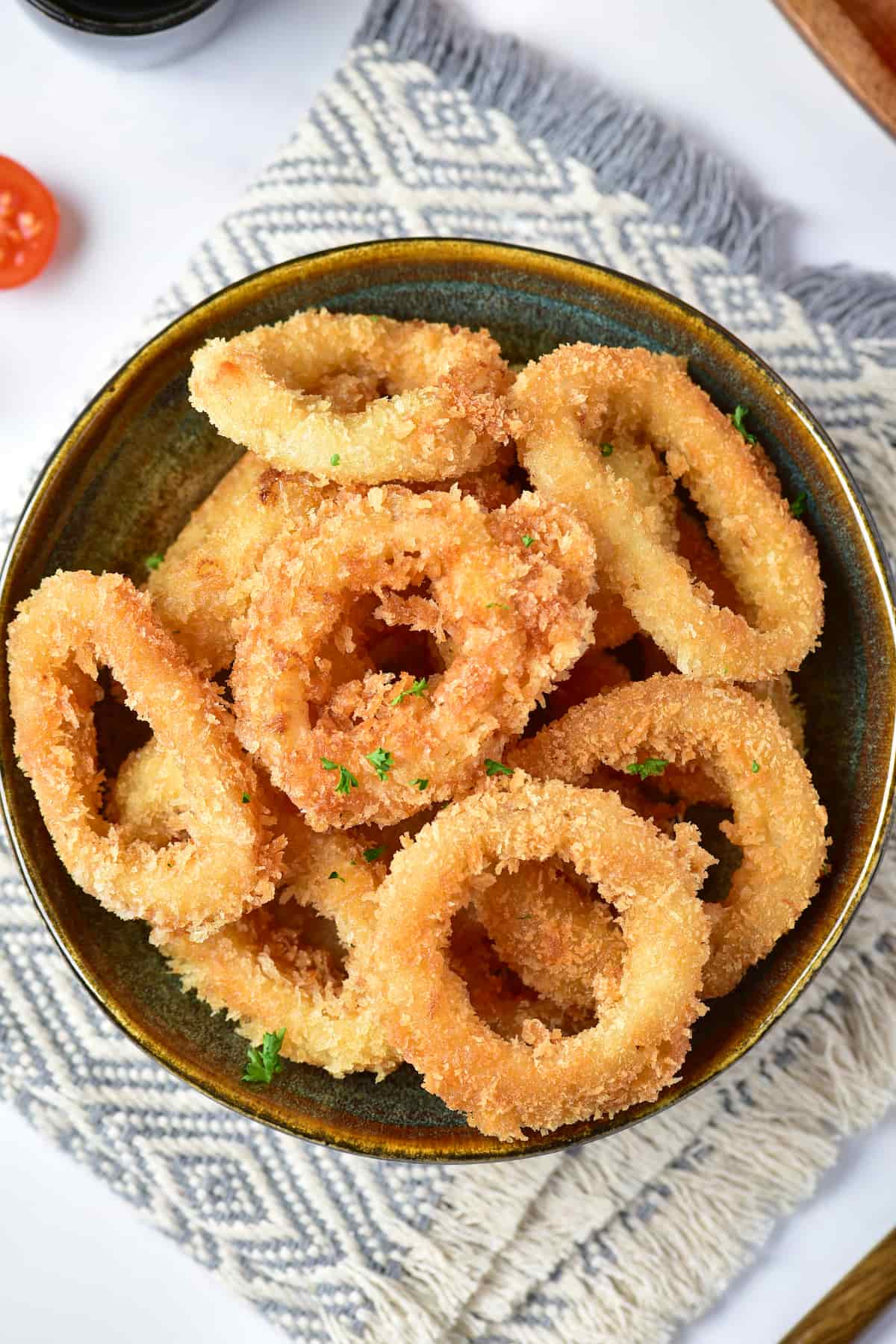Crispy calamari fries in a bowl with sliced lemons and a touch of sauce on one piece, placed on a mat with sliced tomato nearby.