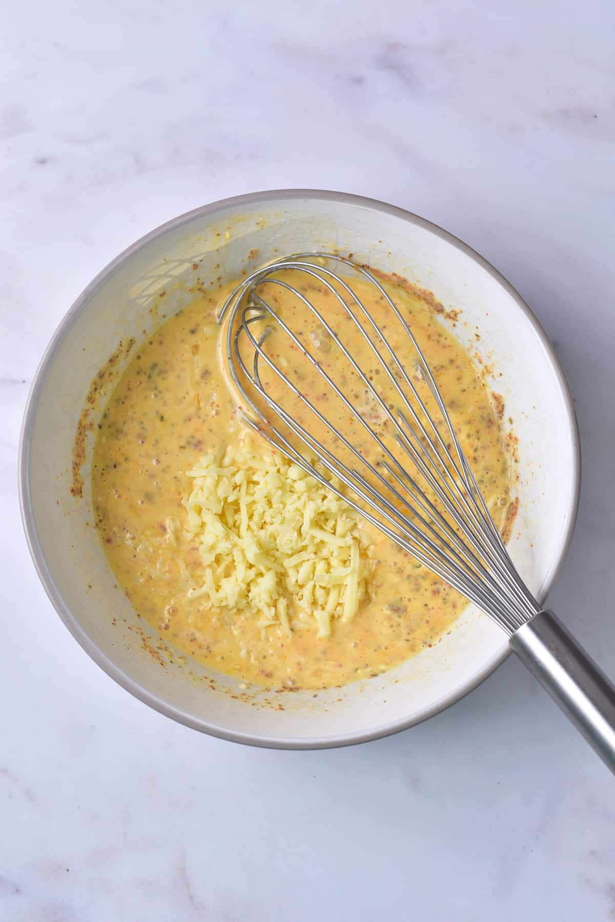 A metal whisk rests in a bowl of beaten eggs mixed with grated cheese on a white marble surface.