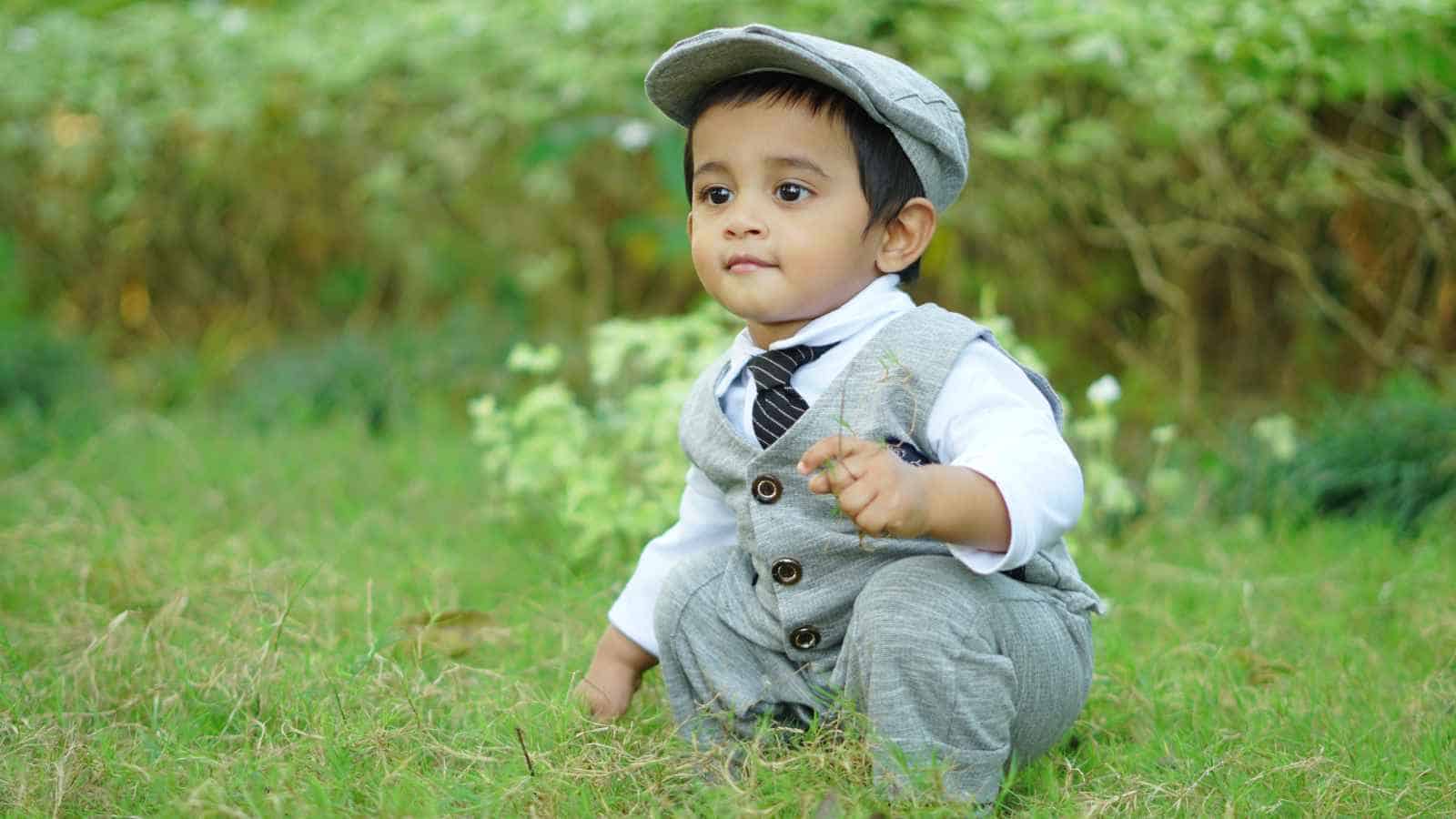 A young child wearing a grey suit, tie, and cap is squatting on grass outdoors, with greenery visible in the background.