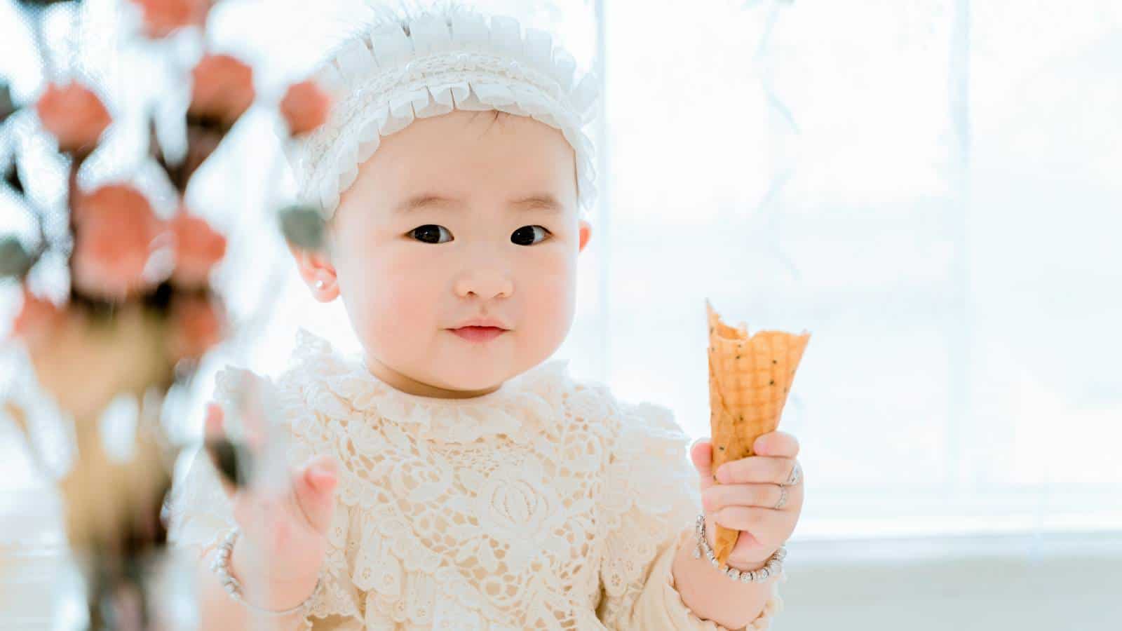A baby wearing a white lace outfit and headband holds an empty ice cream cone, with blurred flowers in the foreground and a bright background.