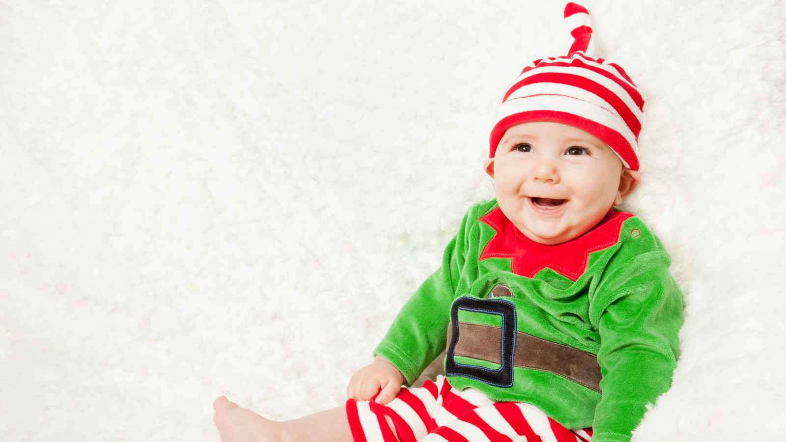 A smiling baby sits on a soft white surface, dressed in a green elf costume with a red-and-white striped hat and matching pants.